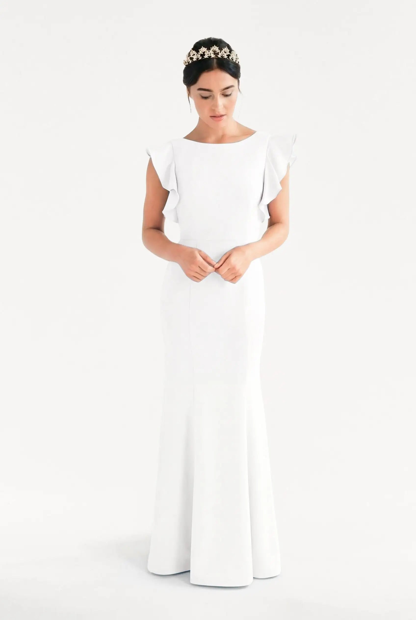 A woman stands against a white background wearing the TH&TH Cecelia Moss Crepe in Ivory—a sleek, floor-length modern bridesmaid dress with ruffled sleeves. She looks down, her dark hair adorned with a decorative floral headband.