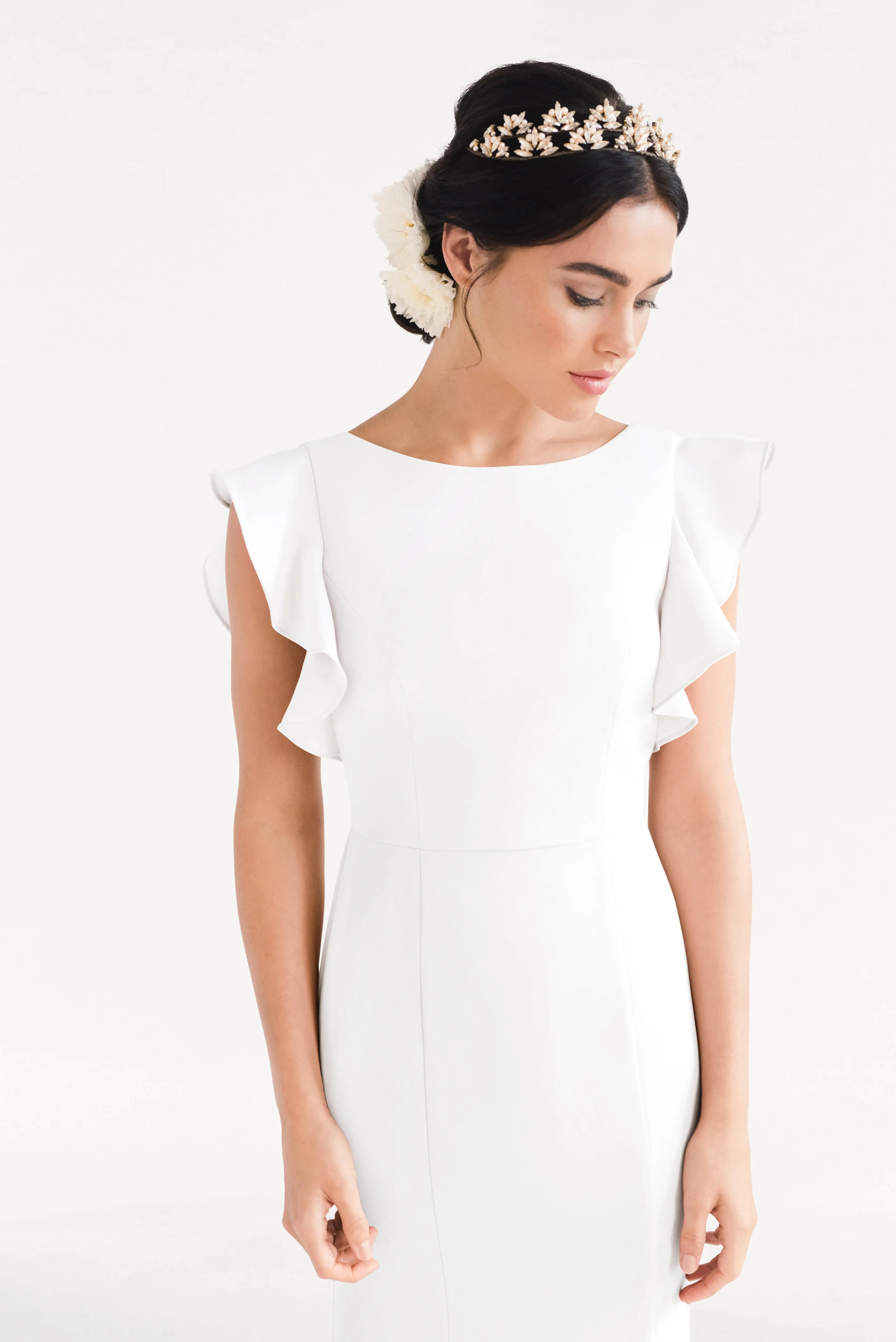 A woman in the TH&TH Cecelia Moss Crepe in Ivory bridesmaid dress stands against a white background. The modern, size-inclusive gown features ruffled shoulders; her dark hair is styled up with a floral headpiece and white accessory.