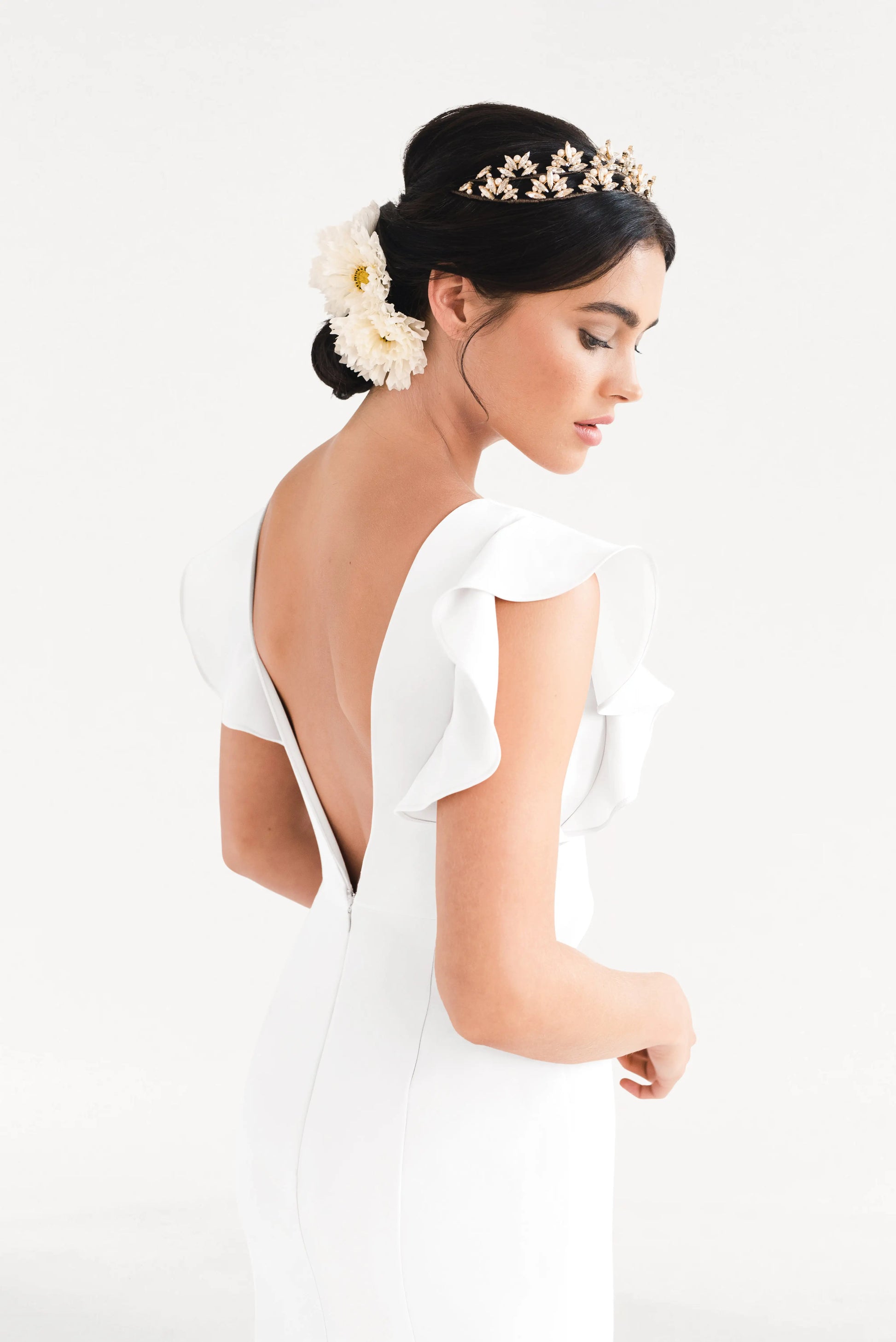 A woman stands in profile wearing the TH&TH Cecelia Moss Crepe in Ivory—a size-inclusive bridesmaid gown with ruffled sleeves and a deep V-back. Her dark hair is styled in a low bun with white flowers and a gold headpiece, set against a white background.