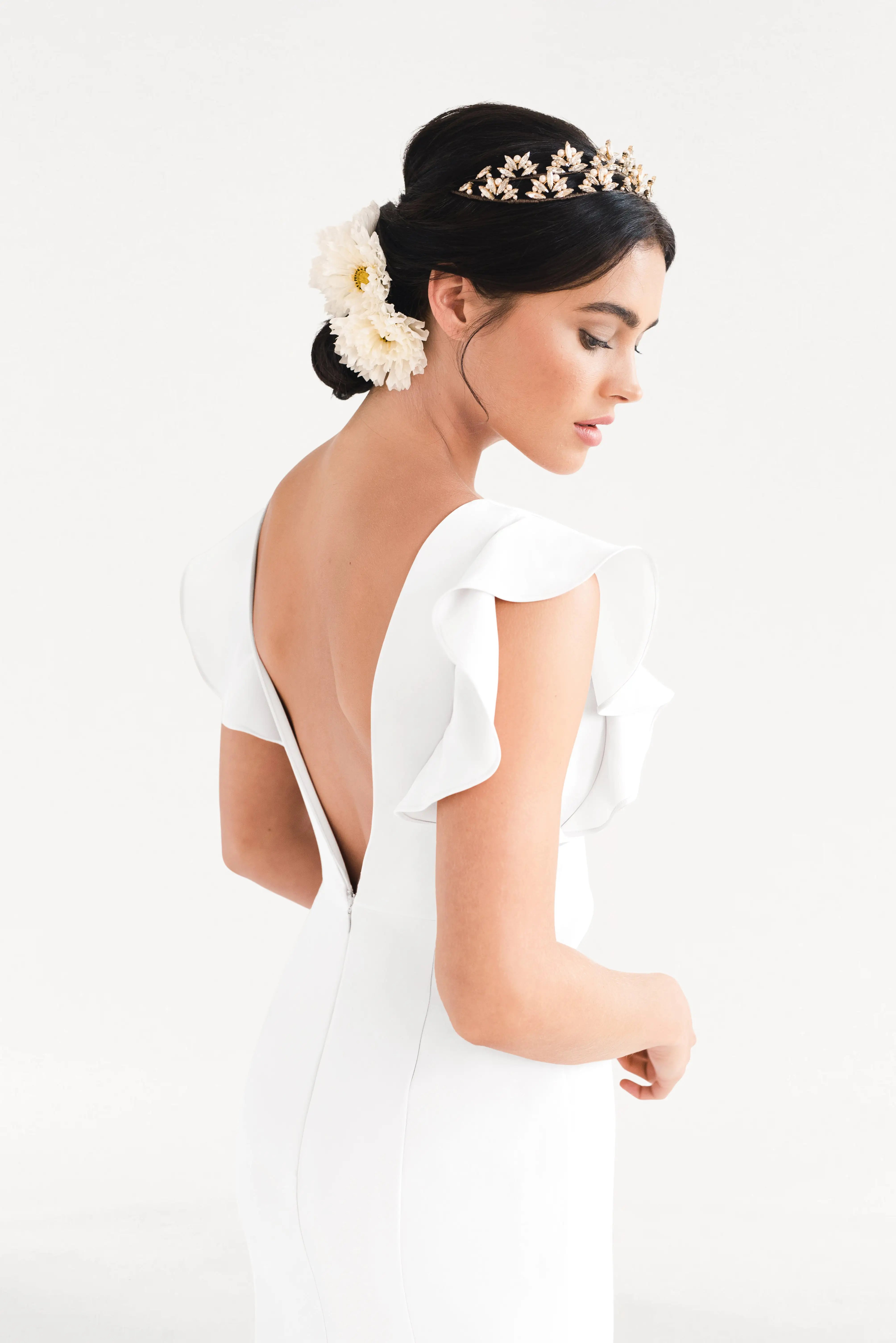 A woman stands in profile wearing the TH&TH Cecelia Moss Crepe in Ivory—a size-inclusive bridesmaid gown with ruffled sleeves and a deep V-back. Her dark hair is styled in a low bun with white flowers and a gold headpiece, set against a white background.