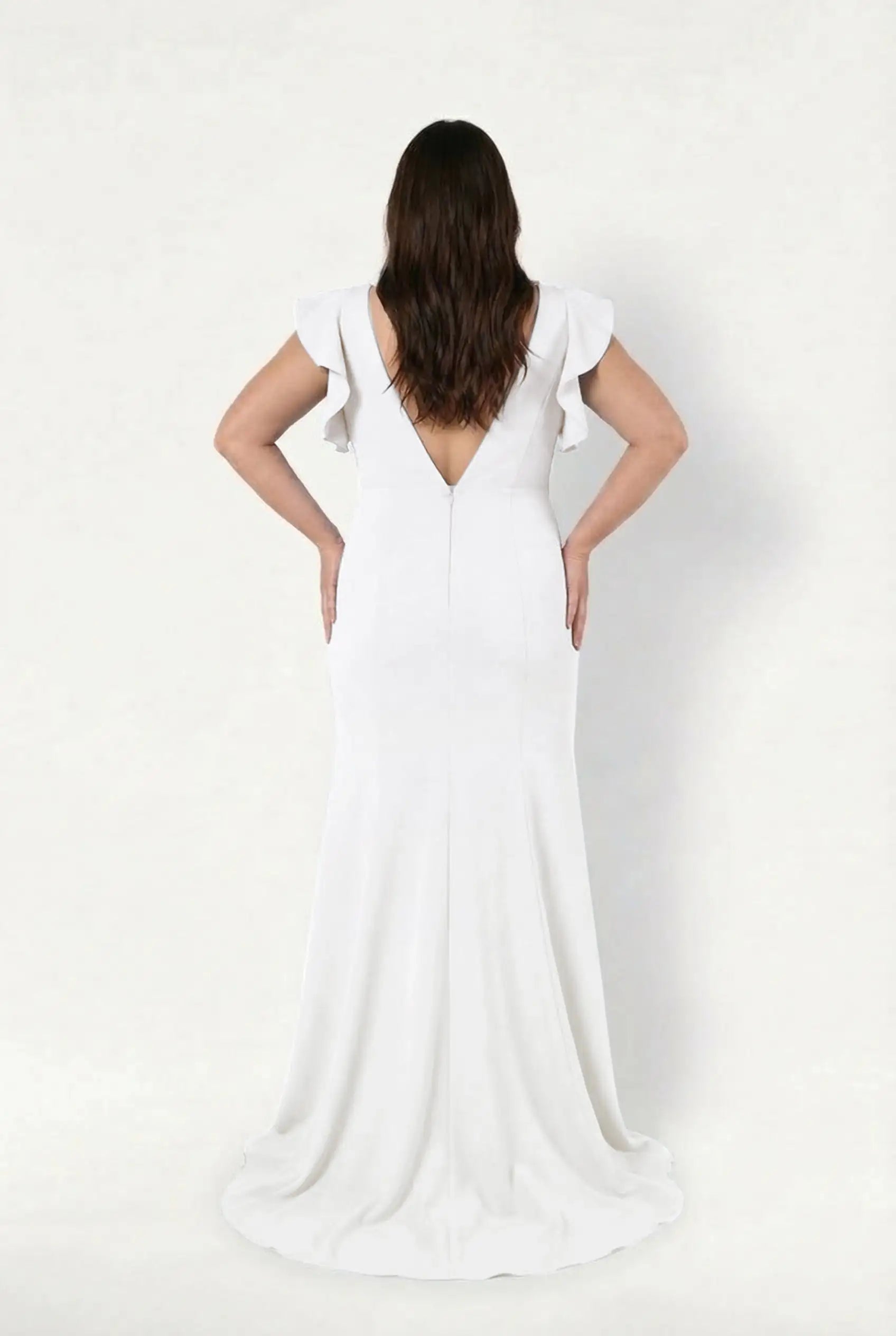 A woman stands facing away in the Cecelia Moss Crepe in Ivory by TH&TH, featuring a deep V-back and ruffled sleeves. Her wavy brown hair cascades over her shoulders as she poses with hands on hips against a plain white backdrop.