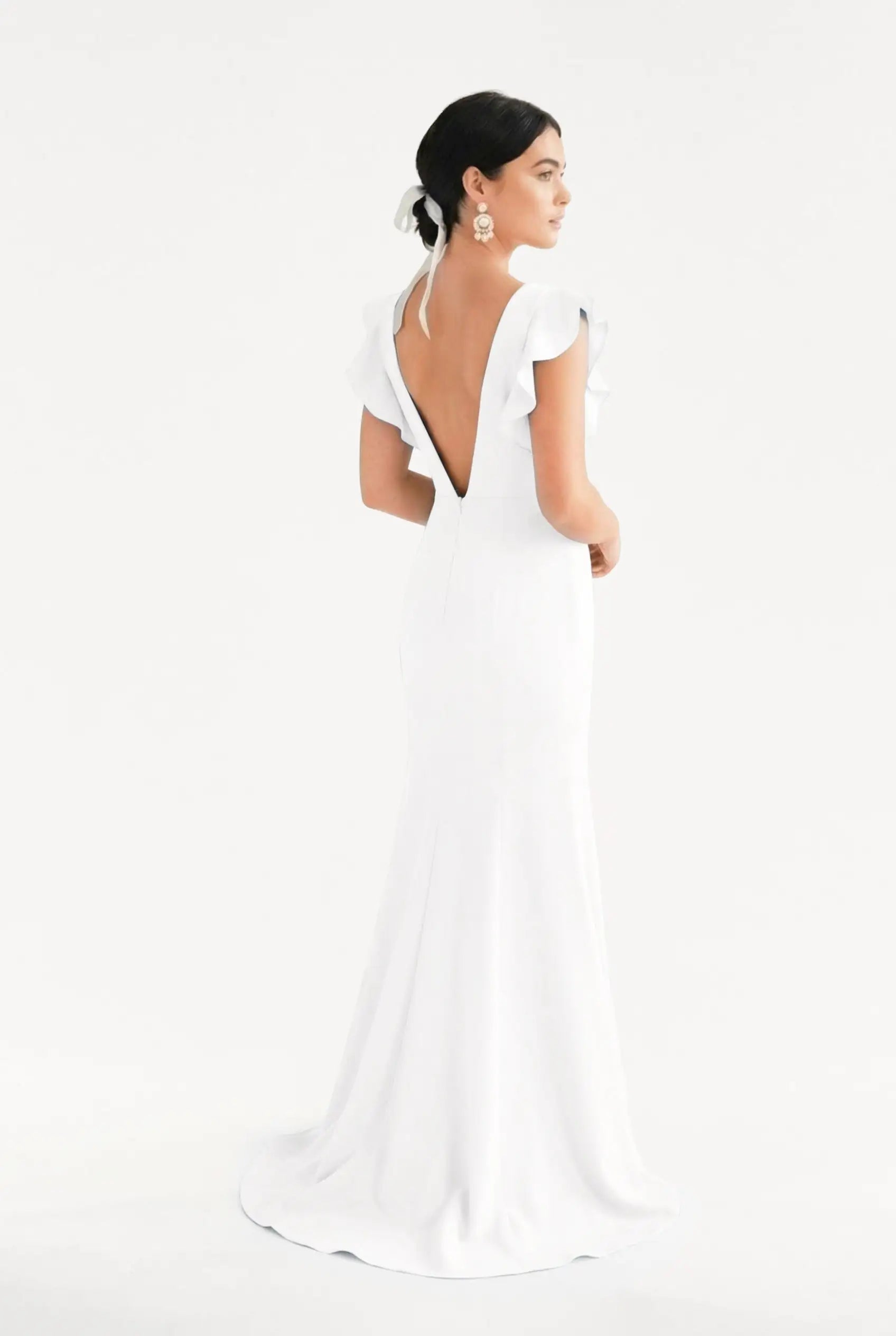A woman faces away in the TH&TH Cecelia Moss Crepe gown in Ivory, featuring ruffled sleeves. Her dark hair is up and she wears bold floral earrings, set against a plain white background.