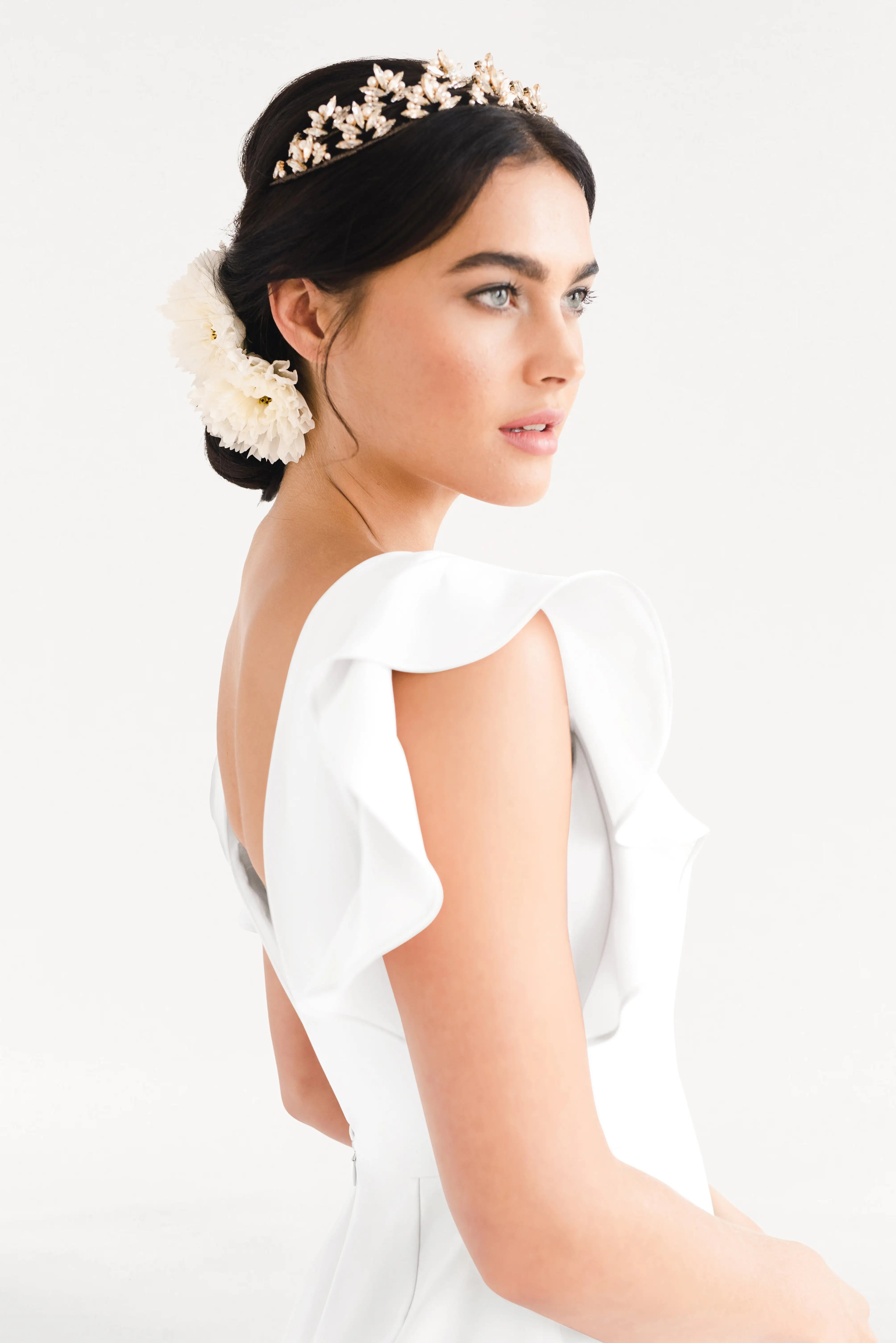 A woman wears the TH&TH Cecelia Moss Crepe in Ivory, a size-inclusive bridesmaid gown with ruffled sleeves. Her dark hair is styled in an updo with a floral headpiece and white flower as she poses thoughtfully against a plain background.