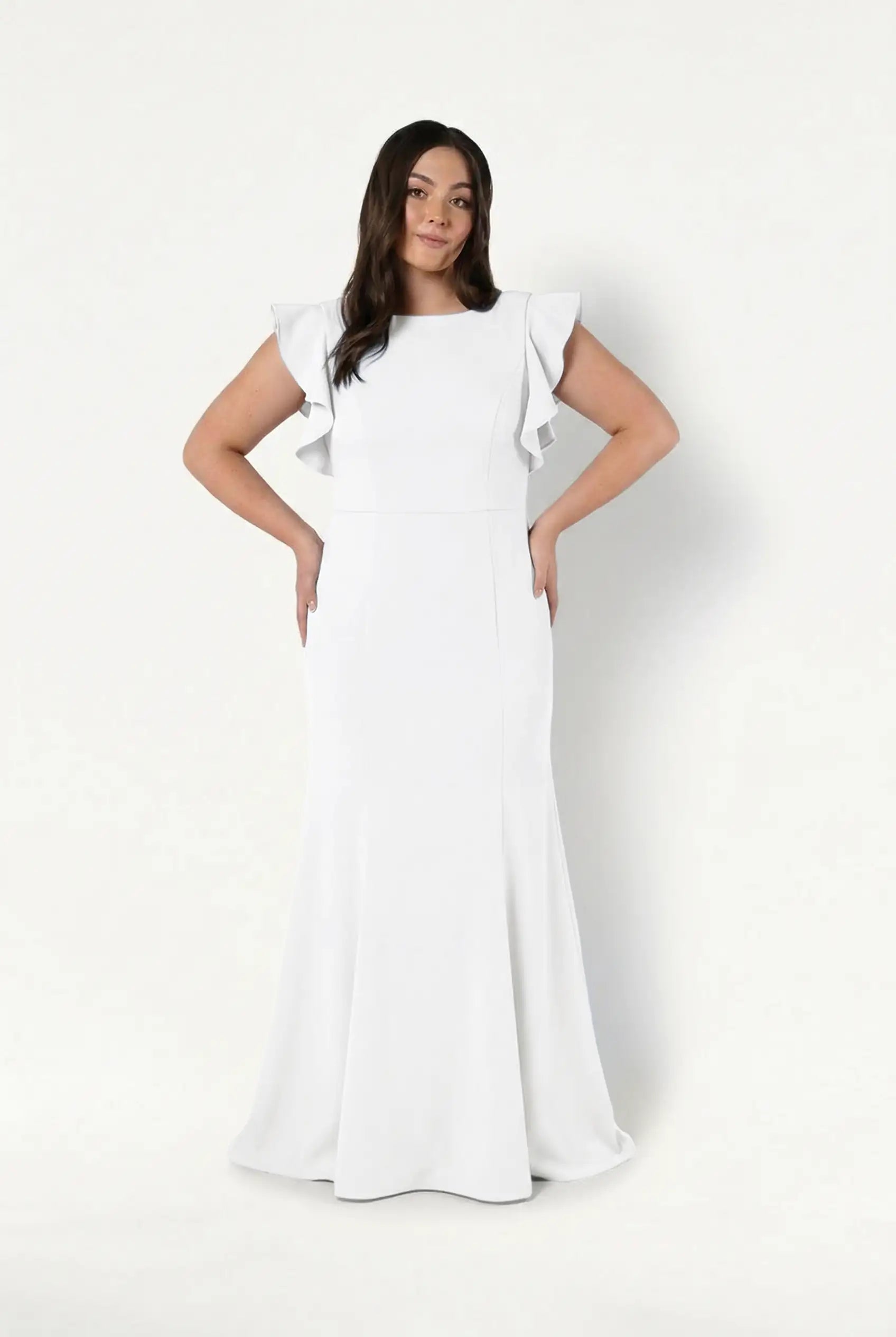 A woman wears the TH&TH Cecelia Moss Crepe in Ivory bridesmaid dress, featuring ruffled cap sleeves and a long, fitted silhouette, standing against a plain white background with her hands on her hips and a slight smile.