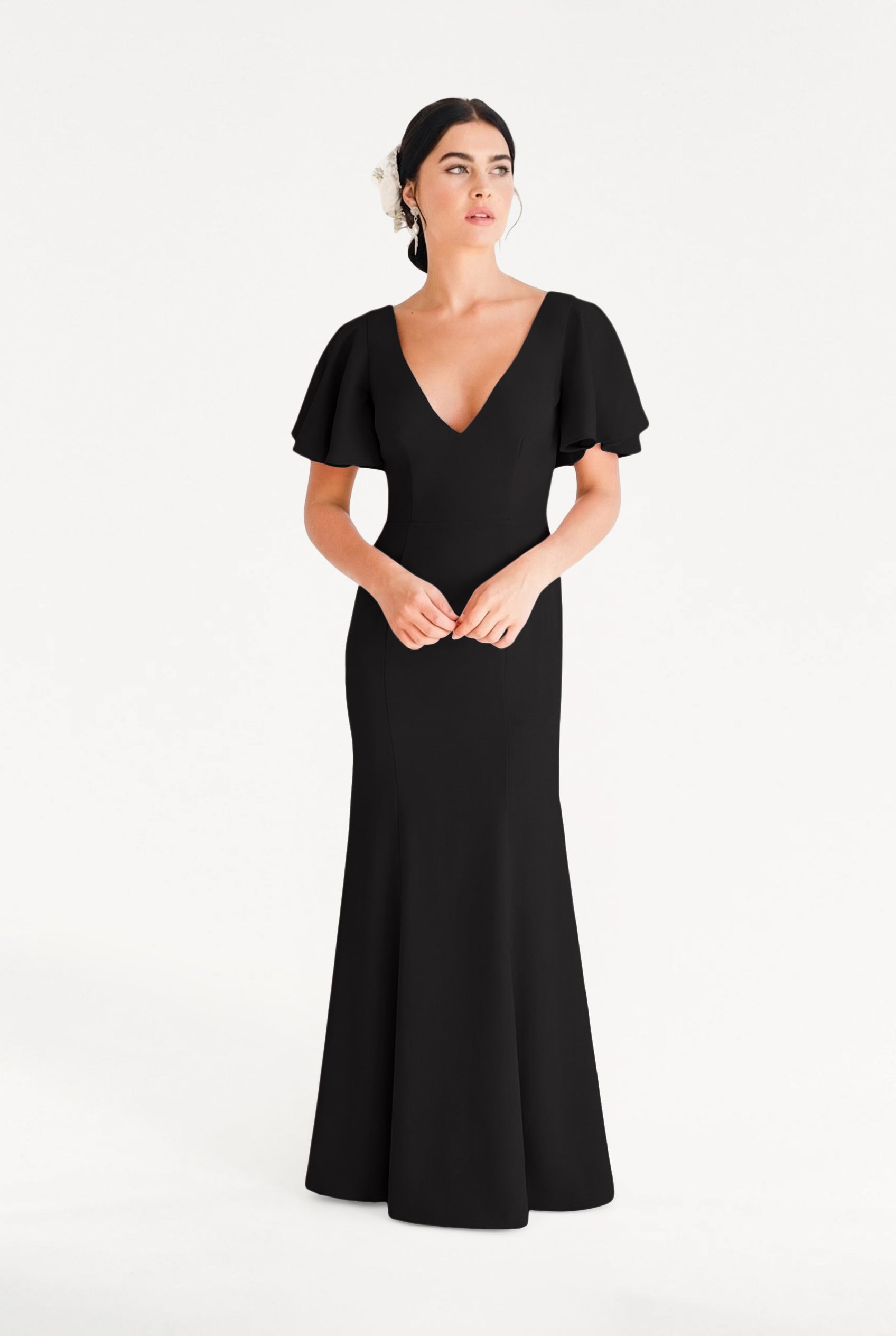 A woman with dark hair styled back with a white floral accessory wears the TH&TH Celeste Moss Crepe Bridesmaid Dress in Black—floor-length, V-neck, short flutter sleeves—standing against a plain white background.