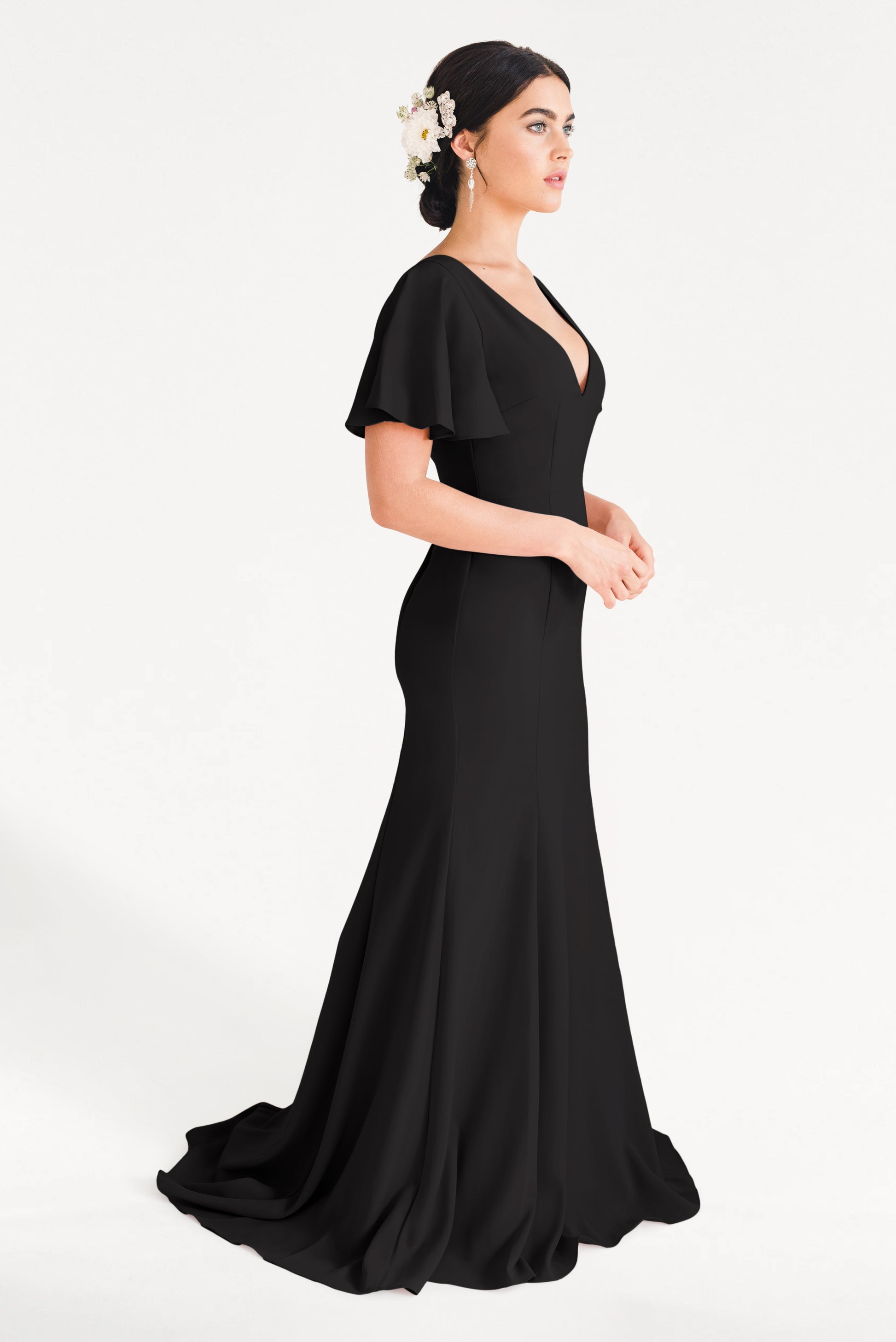 A woman stands in profile wearing the TH&TH Celeste Moss Crepe Bridesmaid Dress in Black, featuring flutter sleeves. Her updo, adorned with white flowers, and drop earrings complete the look against a plain white background.