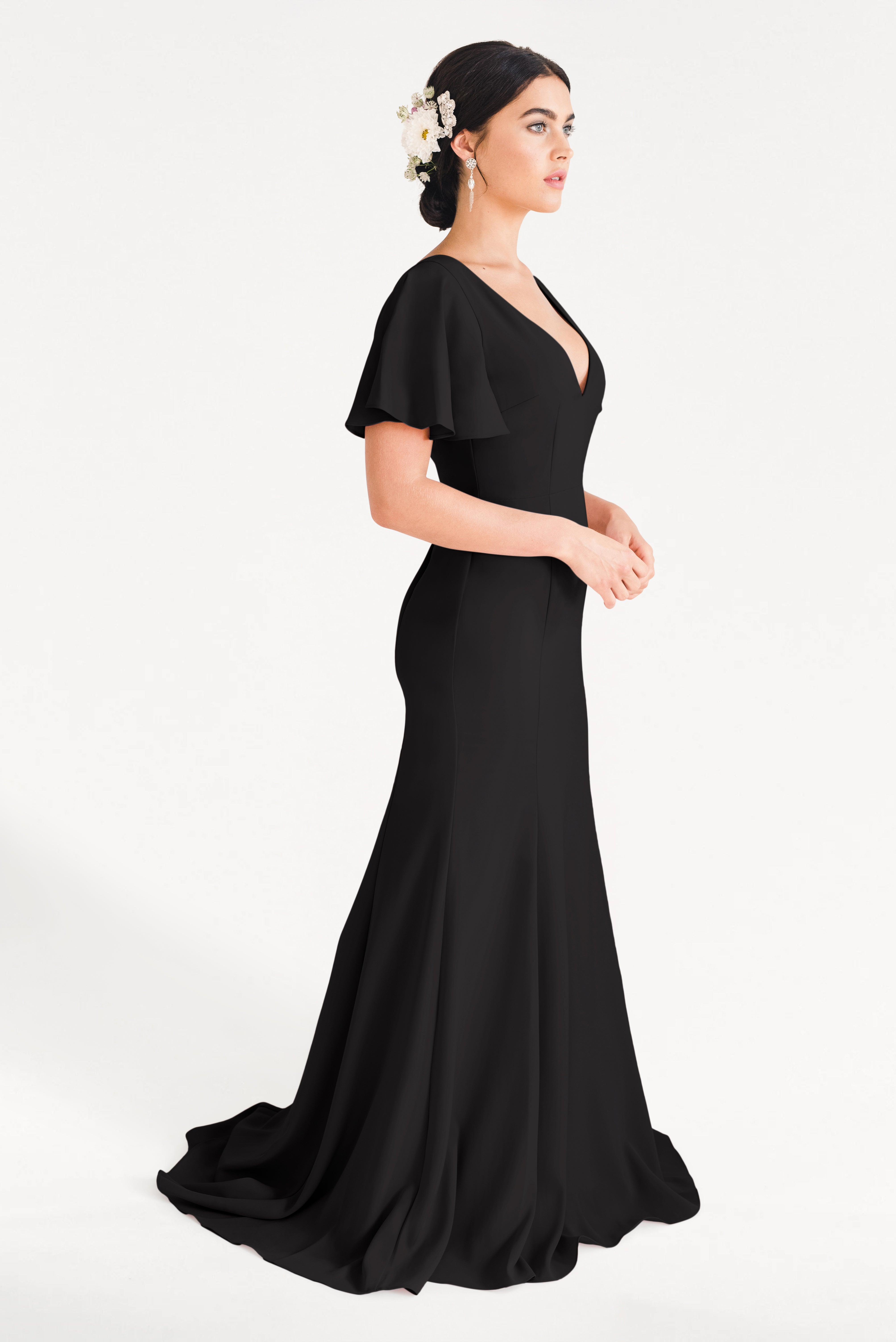 A woman stands in profile wearing the TH&TH Celeste Moss Crepe Bridesmaid Dress in Black, featuring flutter sleeves. Her updo, adorned with white flowers, and drop earrings complete the look against a plain white background.