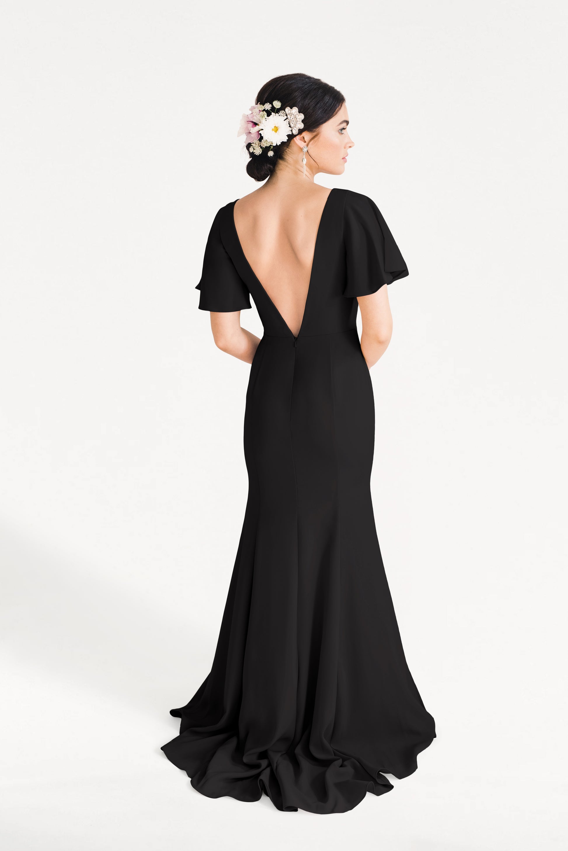 A woman wears the Celeste Moss Crepe Bridesmaid Dress in Black by TH&TH, featuring a long modern cut, deep V-back, and flutter sleeves. Her updo with white and pink flowers enhances the elegant look against a plain white background.
