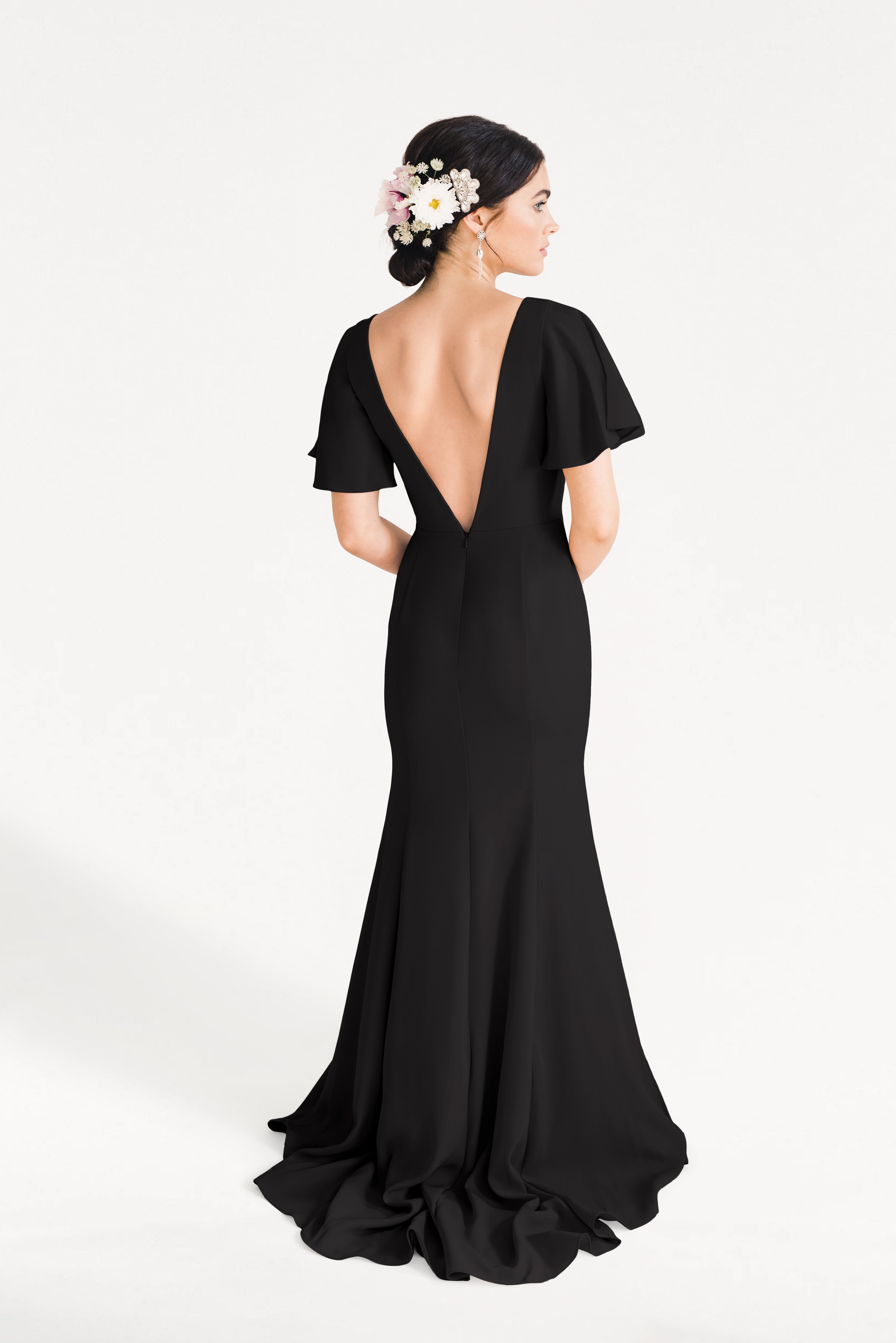 A woman wears the Celeste Moss Crepe Bridesmaid Dress in Black by TH&TH, featuring a long modern cut, deep V-back, and flutter sleeves. Her updo with white and pink flowers enhances the elegant look against a plain white background.