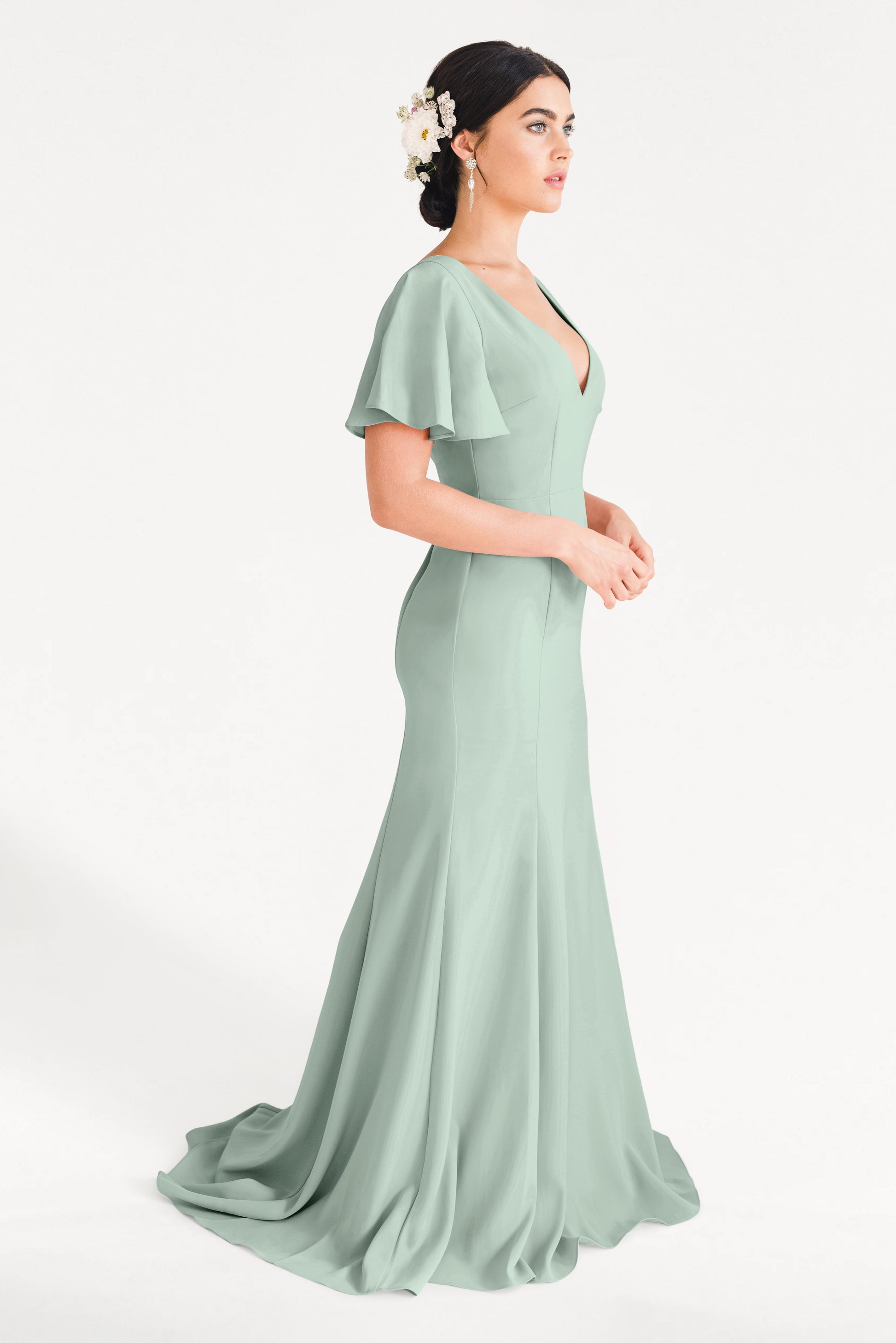 A woman in the TH&TH Celeste Moss Crepe Bridesmaid Dress in Sage Green, a floor-length gown with flutter sleeves, stands in profile. Her dark hair is in an updo with white floral accents and she wears drop earrings against a plain white background.
