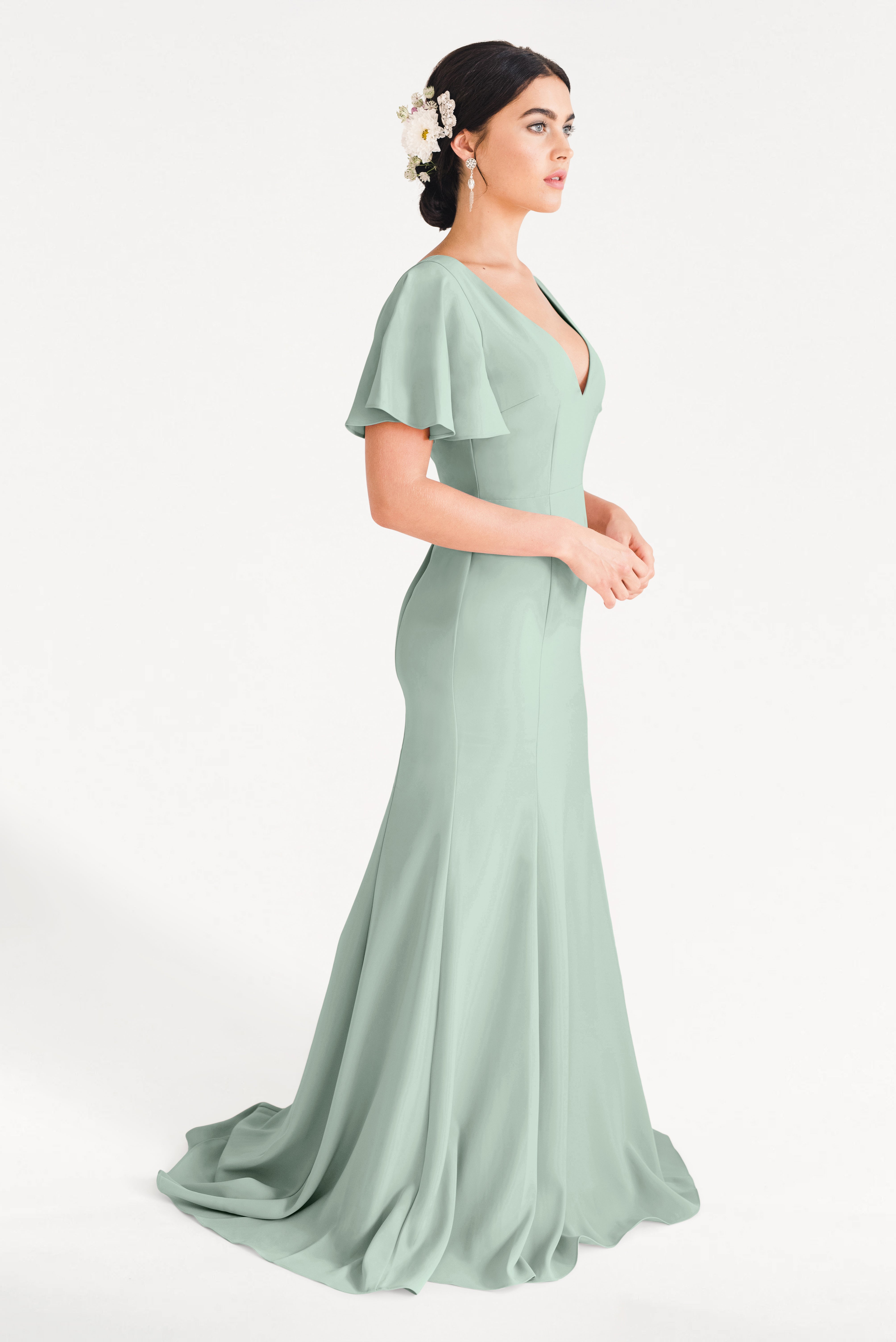 A woman in the TH&TH Celeste Moss Crepe Bridesmaid Dress in Sage Green, a floor-length gown with flutter sleeves, stands in profile. Her dark hair is in an updo with white floral accents and she wears drop earrings against a plain white background.