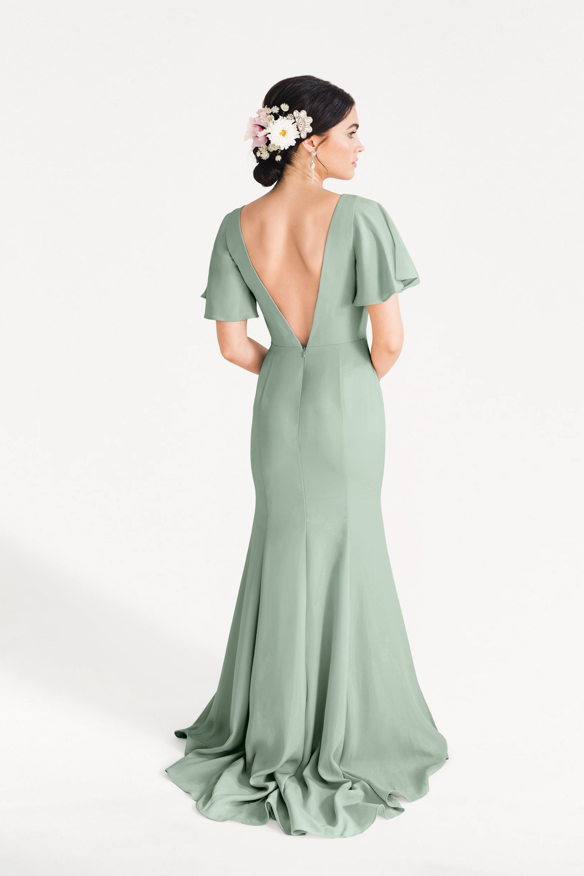 A woman with dark hair in a floral bun models the Celeste Moss Crepe Bridesmaid Dress in Sage Green by TH&TH—a size-inclusive gown featuring flutter sleeves and a deep V-back—against a white background.