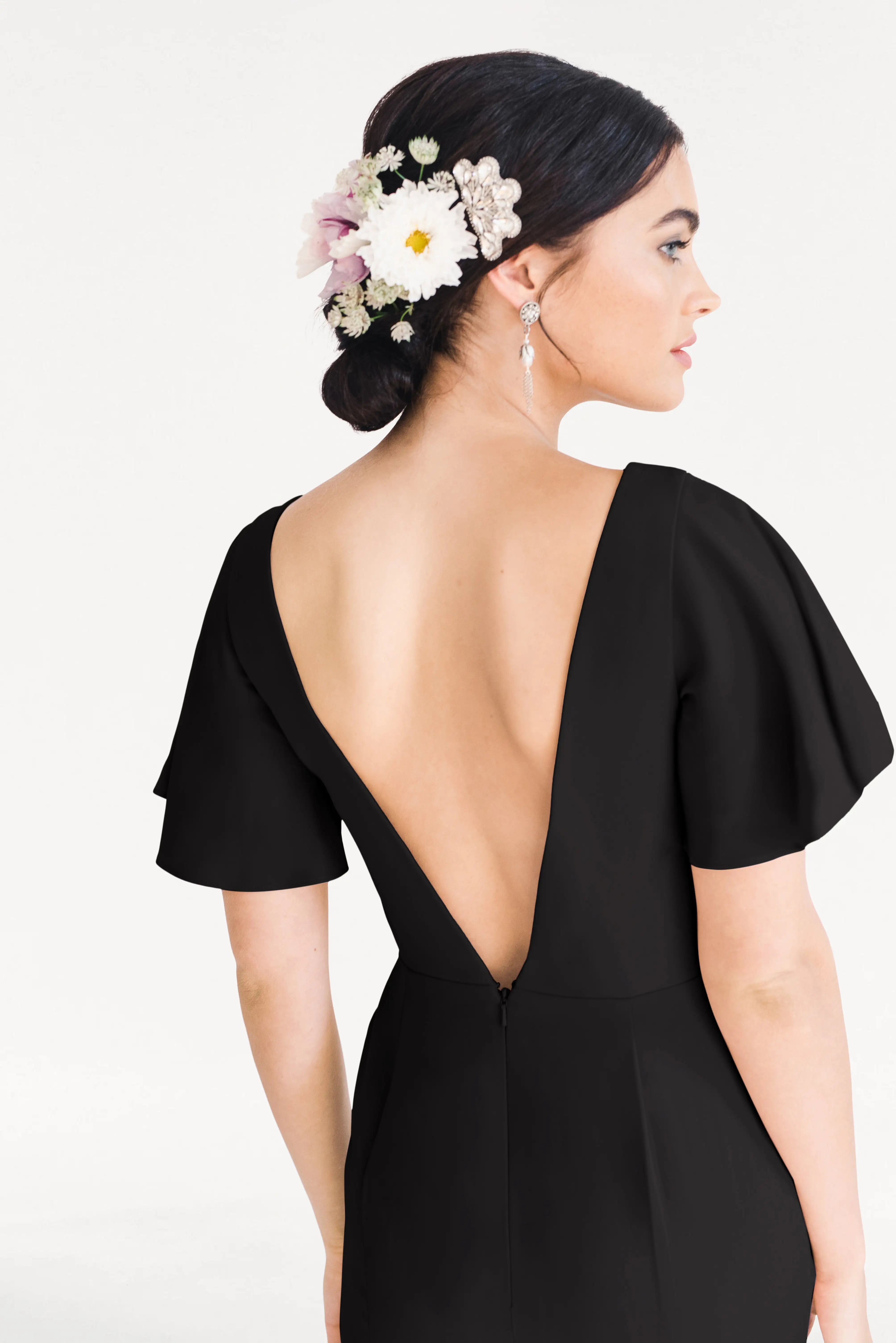 A woman models the TH&TH Celeste Moss Crepe Bridesmaid Dress in Black, featuring a size-inclusive fit and low-cut back. She has dark hair in a floral-accented low bun and wears drop earrings, posed against a plain light background.