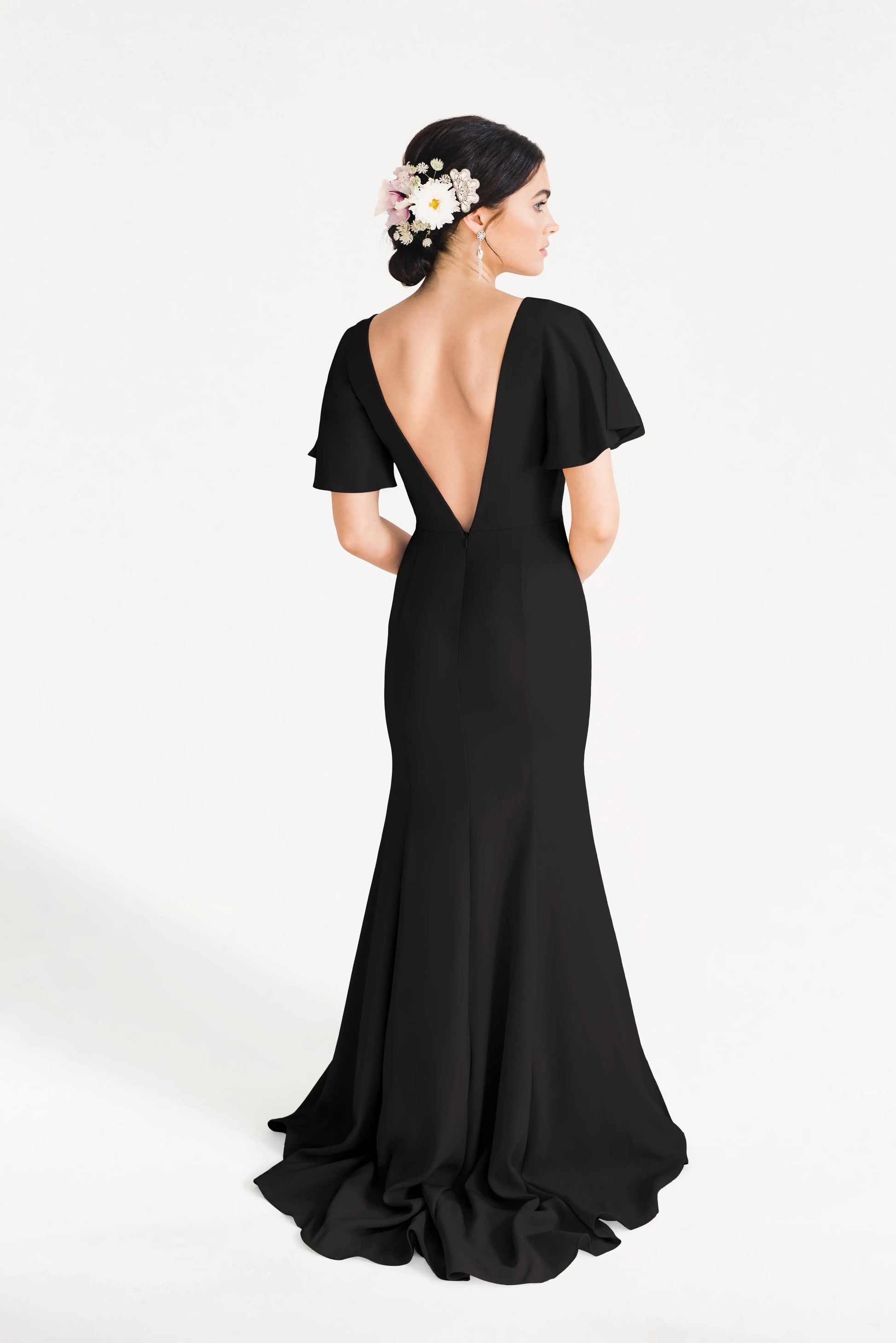 A woman wears the Celeste Moss Crepe Bridesmaid Dress in Black by TH&TH, featuring a long modern cut, deep V-back, and flutter sleeves. Her updo with white and pink flowers enhances the elegant look against a plain white background.