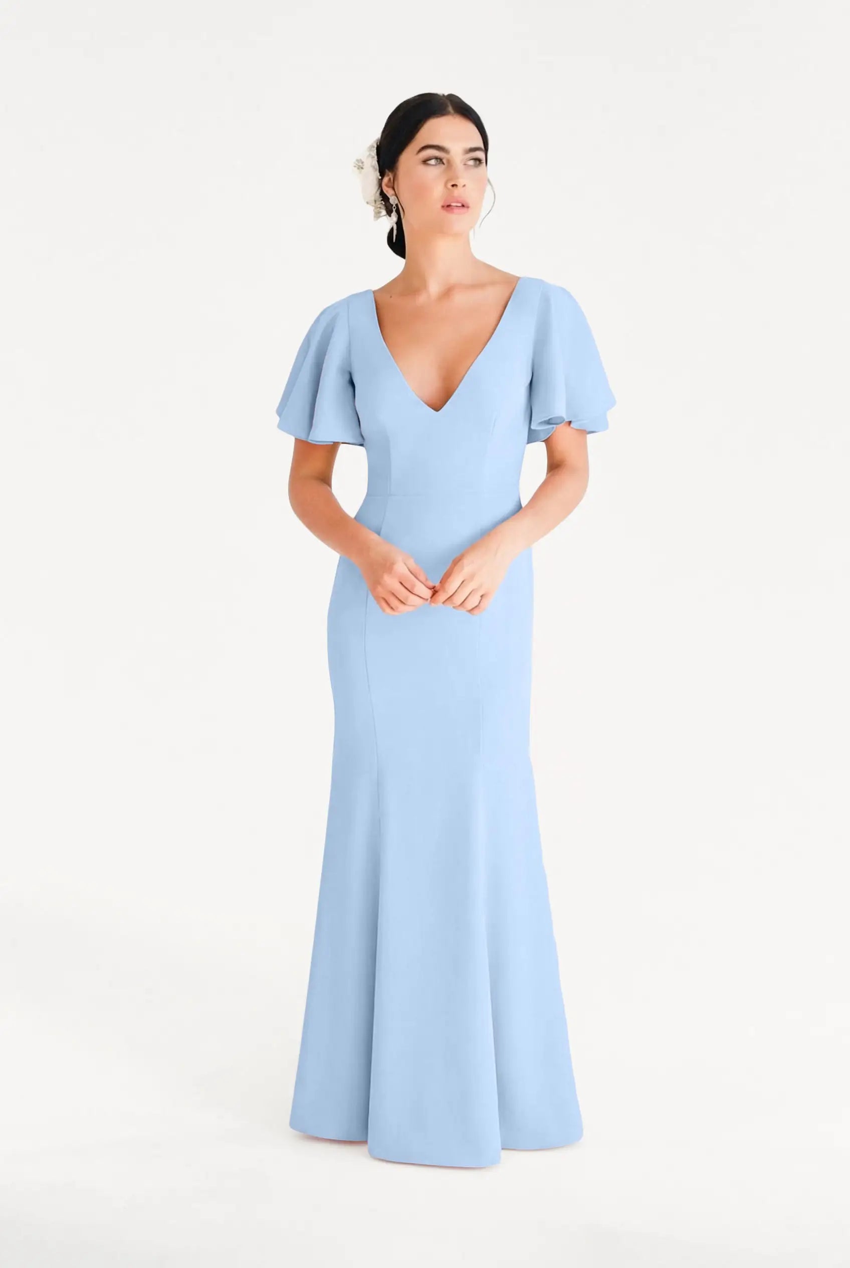 A woman stands against a white background wearing the TH&TH Celeste Moss Crepe Bridesmaid Dress in Dusty Blue, a floor-length modern gown with short flutter sleeves and a deep V-neckline. Her dark hair is styled back with a white flower accessory.