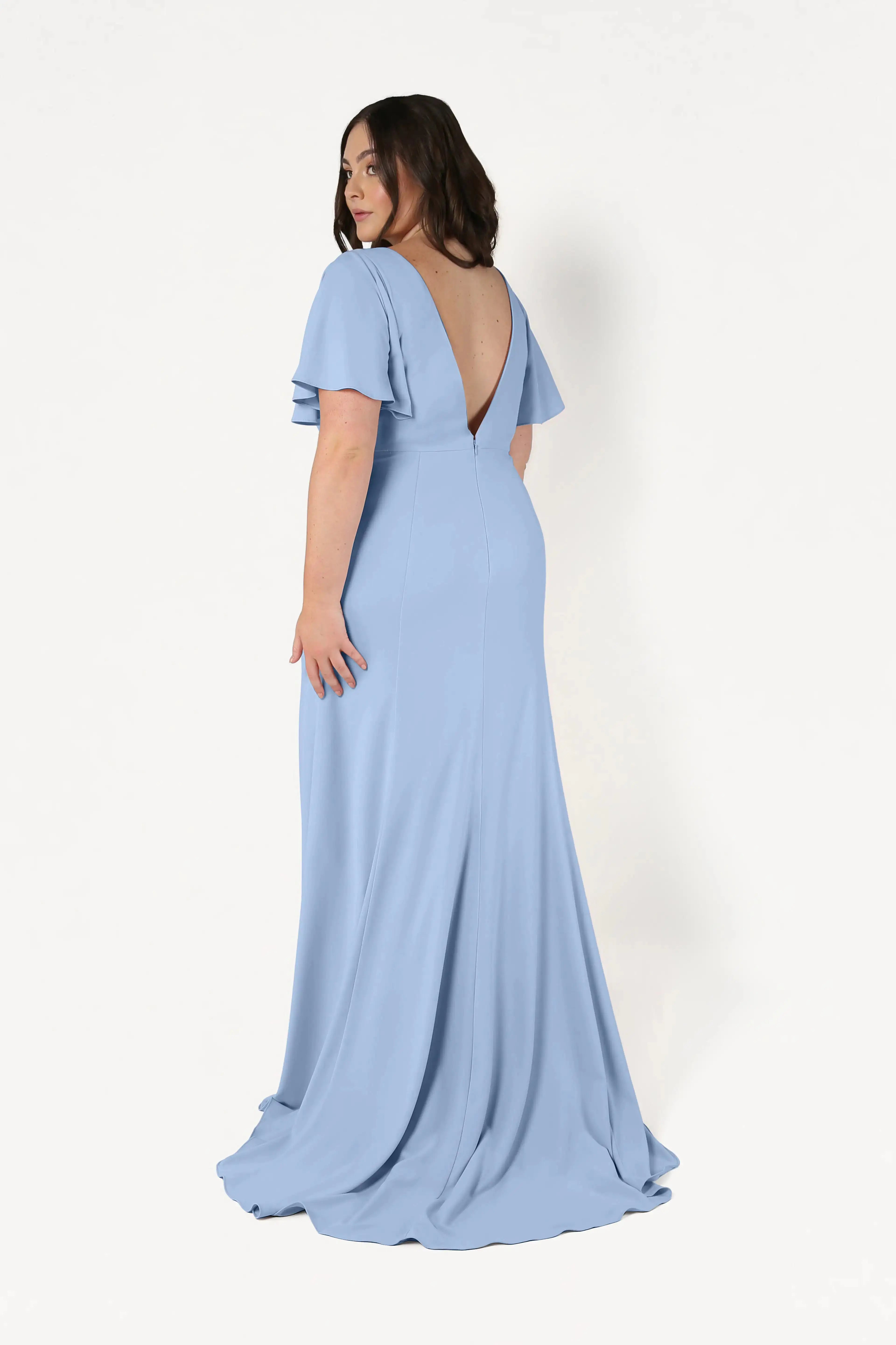 A woman with long brown hair wears the TH&TH Celeste Moss Crepe Bridesmaid Dress in Dusty Blue—a floor-length gown with flutter sleeves and a deep V-shaped back—standing against a plain white background, slightly turned to show the back.