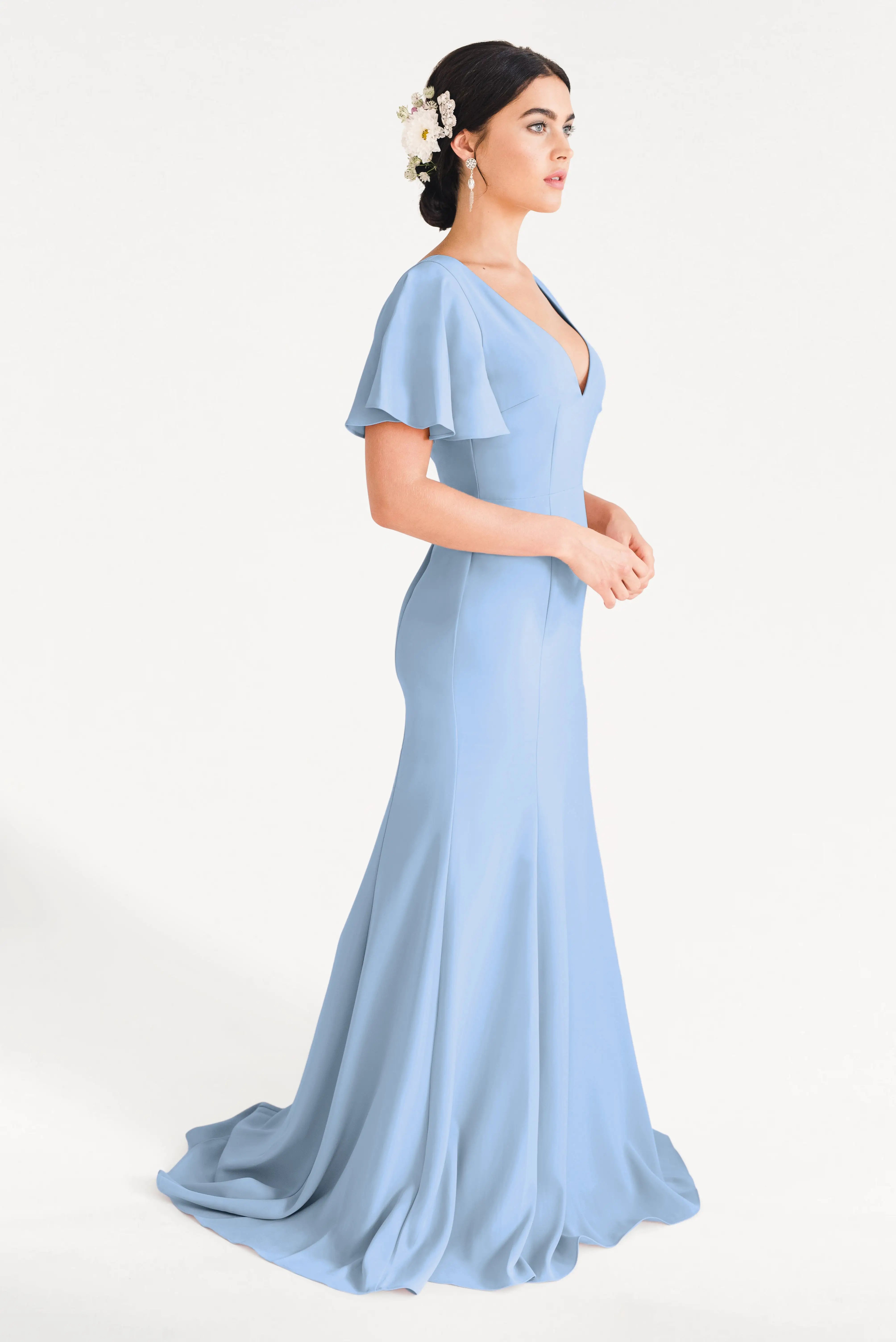 A woman in profile wears the TH&TH Celeste Moss Crepe Bridesmaid Dress in Dusty Blue, featuring a V-neckline. Her dark updo is adorned with white flowers, accentuating the modern elegance of this bridesmaid dress against a white background.