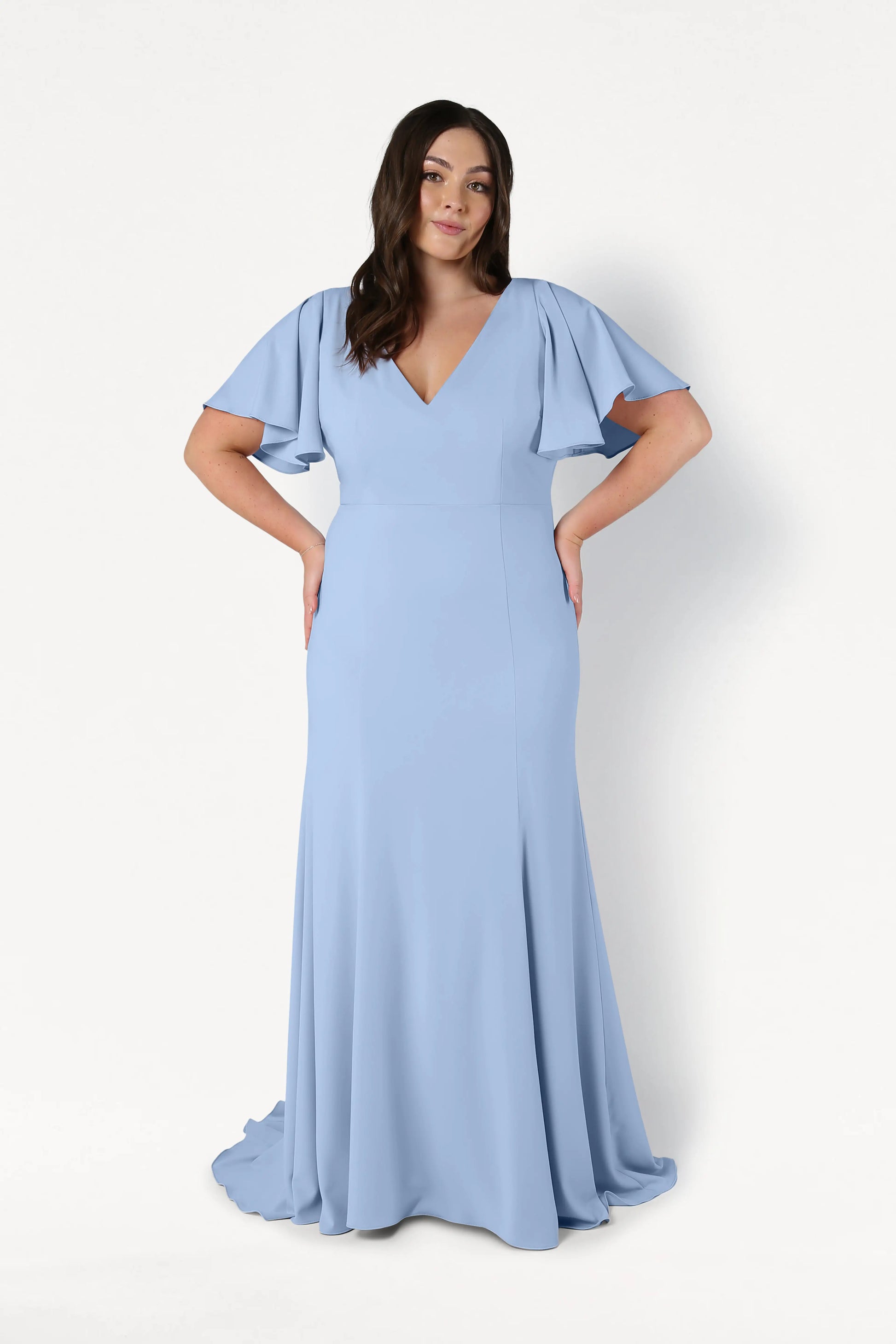 A woman poses against a white background in the TH&TH Celeste Moss Crepe Bridesmaid Dress in Dusty Blue, a floor-length gown featuring short flutter sleeves and a V-neckline. Her hands are on her hips, and her dark hair is styled in loose waves.