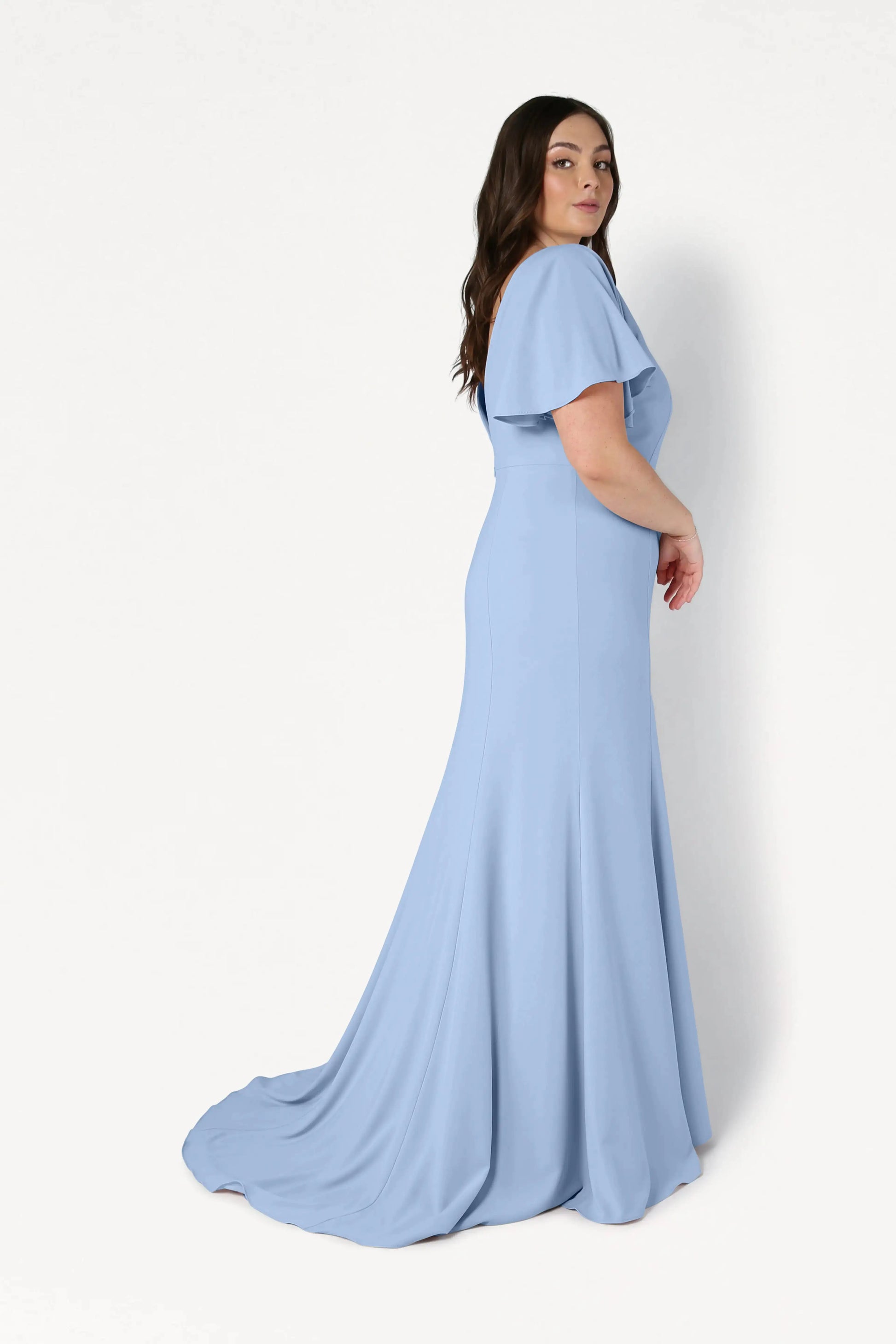 A woman with long dark hair models the size-inclusive TH&TH Celeste Moss Crepe Bridesmaid Dress in Dusty Blue, featuring short flutter sleeves and a slight train, standing sideways against a white background.