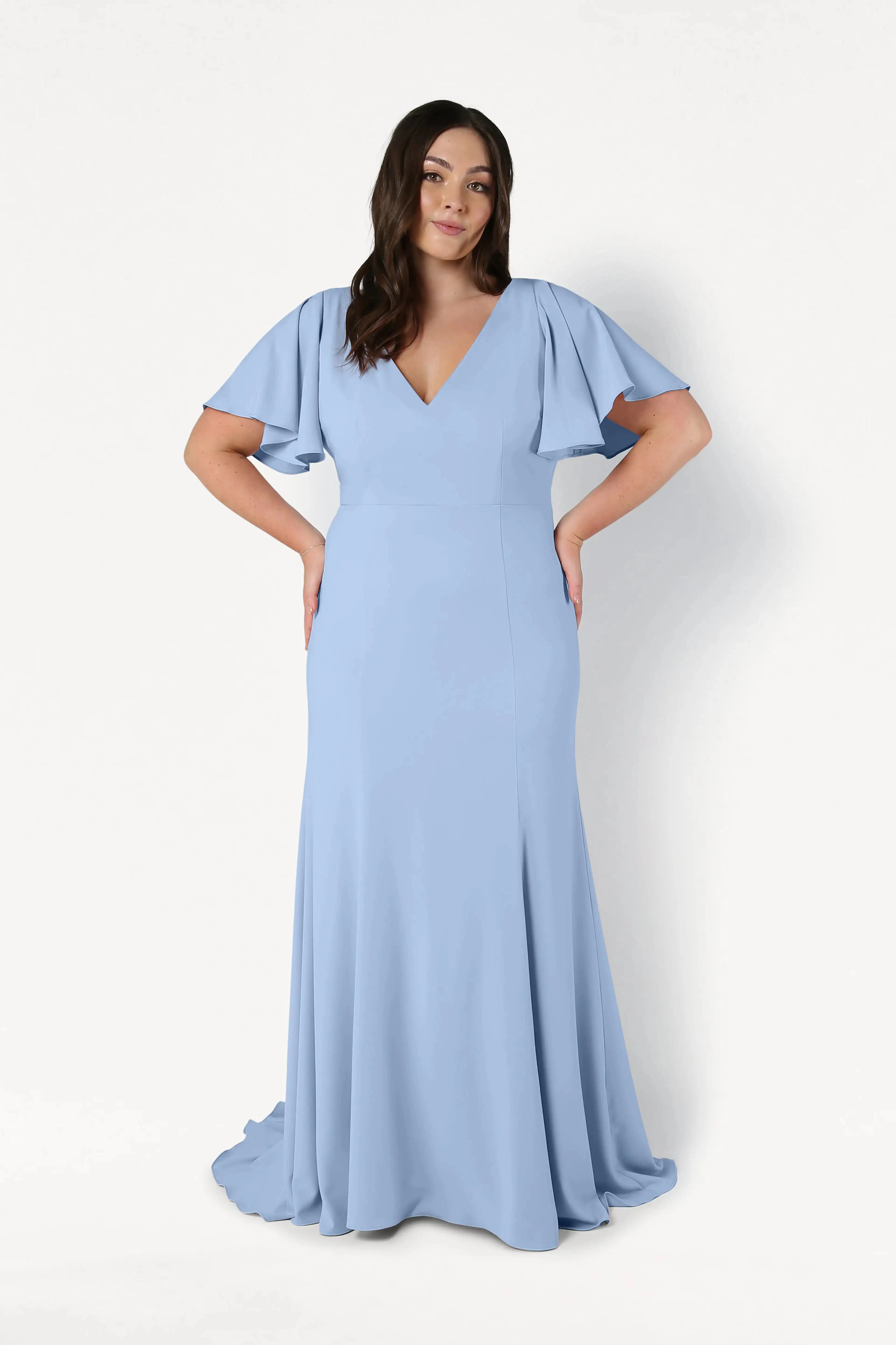 A woman poses against a white background in the TH&TH Celeste Moss Crepe Bridesmaid Dress in Dusty Blue, a floor-length gown featuring short flutter sleeves and a V-neckline. Her hands are on her hips, and her dark hair is styled in loose waves.