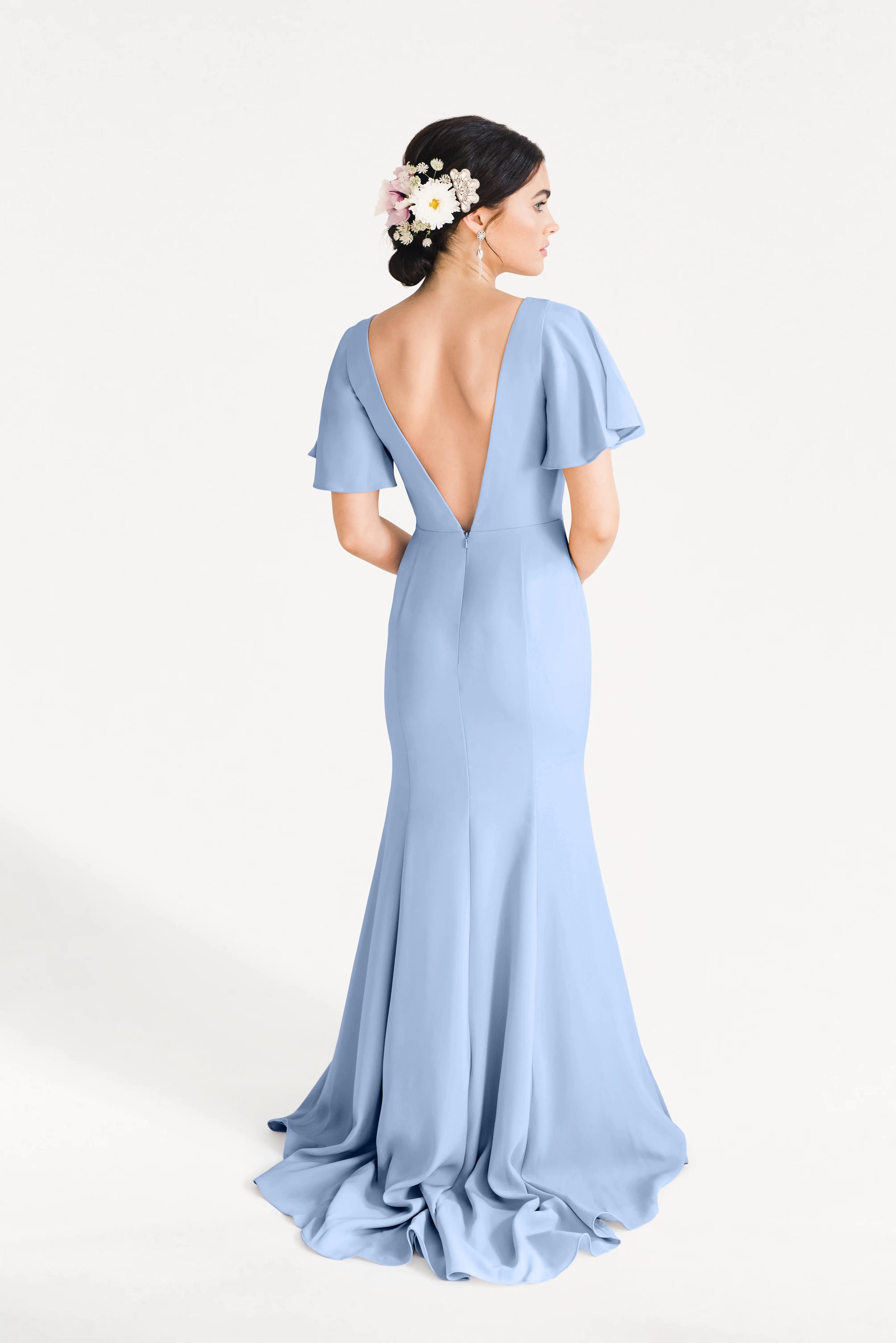 A woman stands facing away in the TH&TH Celeste Moss Crepe Bridesmaid Dress in Dusty Blue, a floor-length gown with flutter sleeves and a deep V-back. Her dark hair is styled in an updo decorated with white and pink flowers.