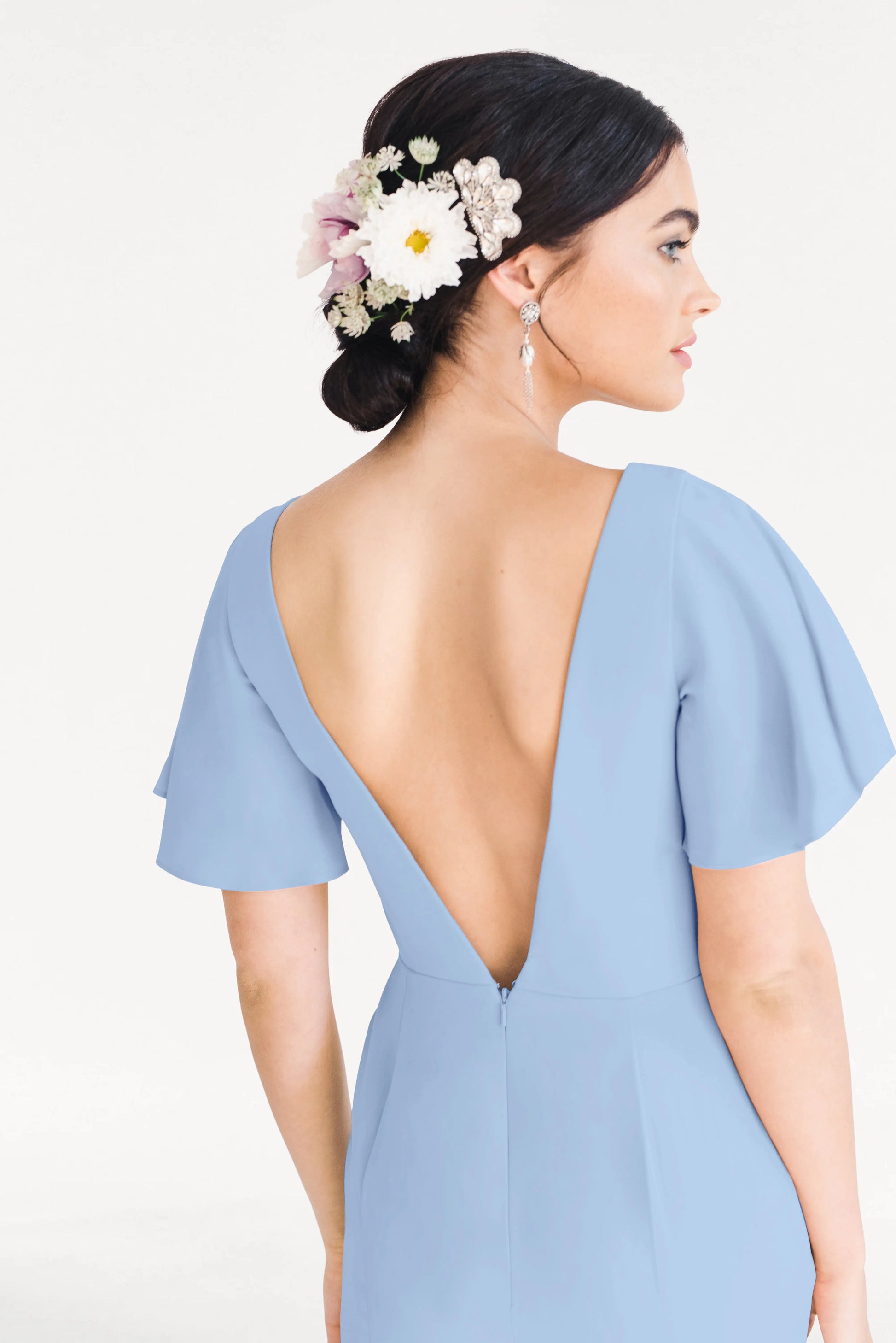A woman with dark hair in a low bun adorned with white and pink flowers models the TH&TH Celeste Moss Crepe Bridesmaid Dress in Dusty Blue, featuring flutter sleeves and a deep V-back, highlighting its elegant, size-inclusive design.