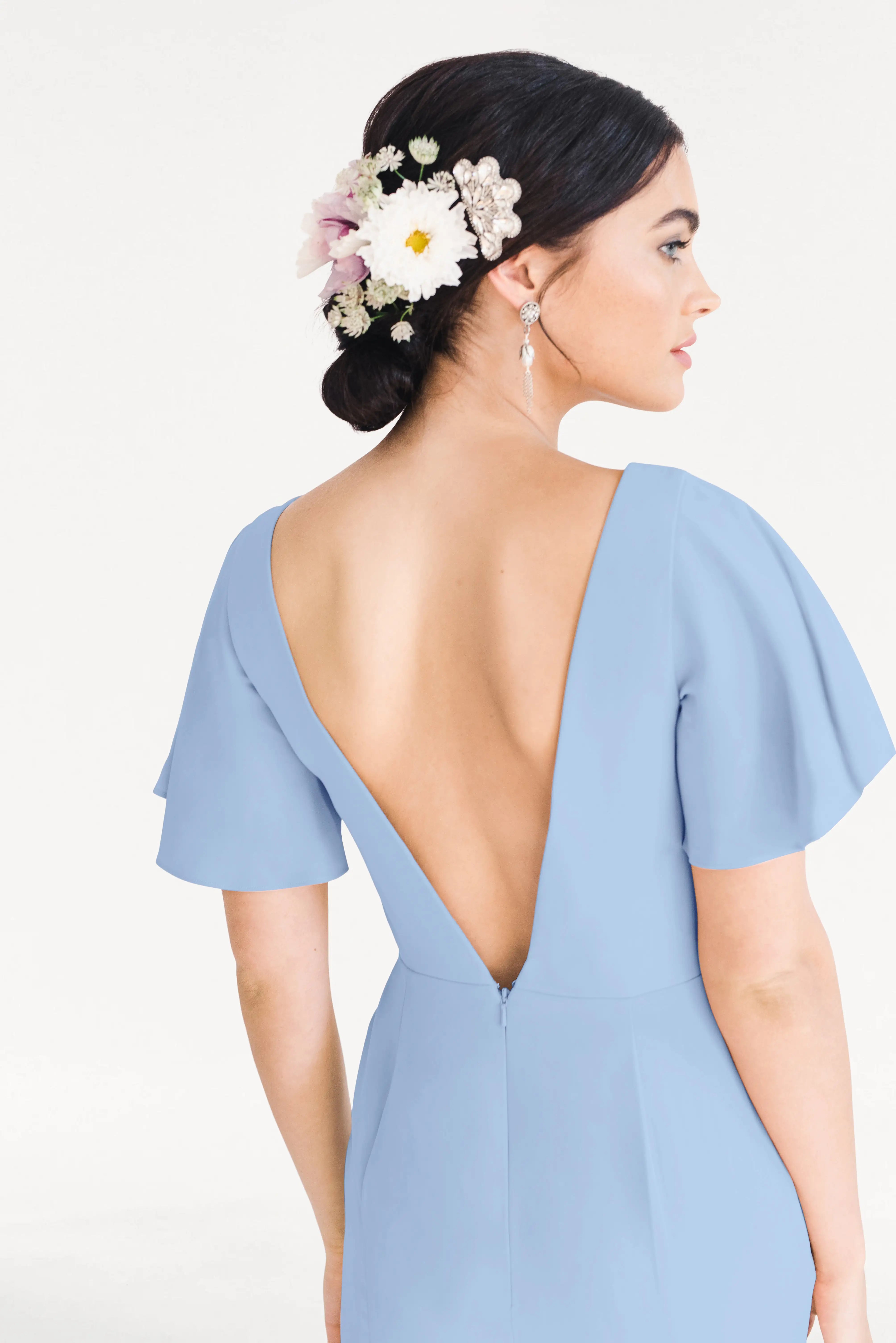 A woman with dark hair in a low bun adorned with white and pink flowers models the TH&TH Celeste Moss Crepe Bridesmaid Dress in Dusty Blue, featuring flutter sleeves and a deep V-back, highlighting its elegant, size-inclusive design.