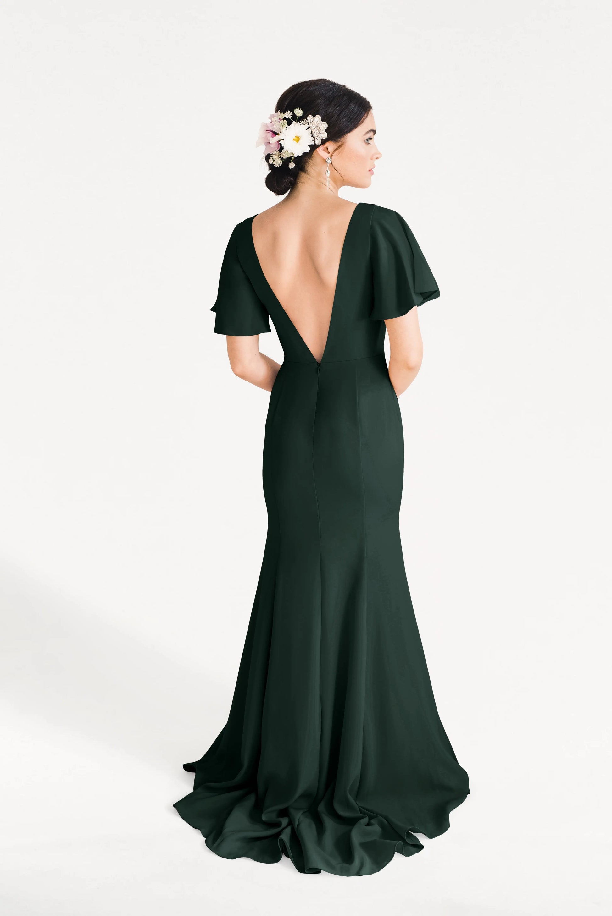 A woman stands with her back to the camera in the fitted, floor-length Celeste Moss Crepe Bridesmaid Dress in Forest Green by TH&TH, featuring flutter sleeves and a deep V-back. Her dark hair is styled up with white and pink flowers against a plain white background.