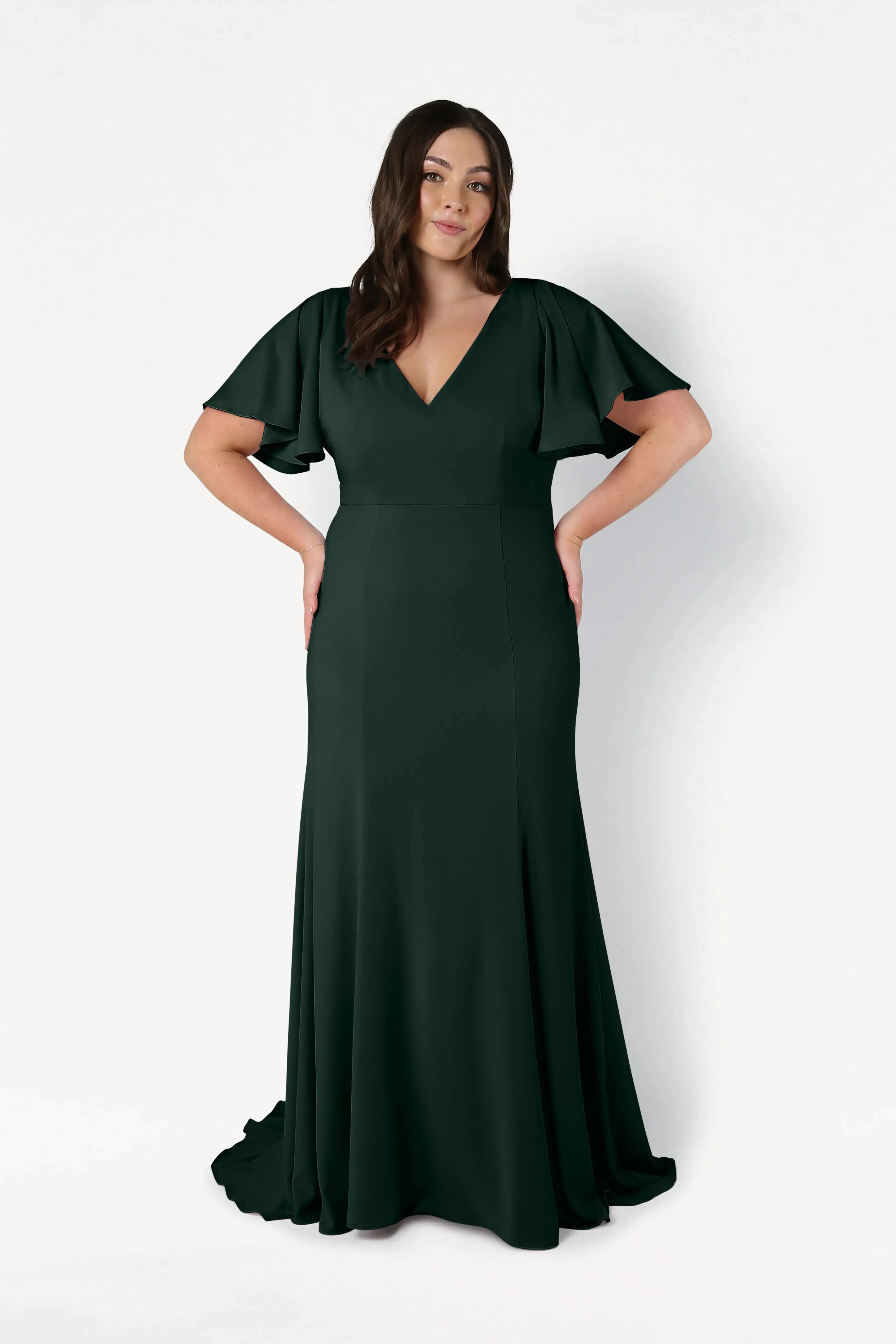 A woman poses against a white background in TH&TH's Celeste Moss Crepe Bridesmaid Dress in Forest Green, featuring flutter sleeves and a V-neckline—an elegant, modern choice for bridal parties.