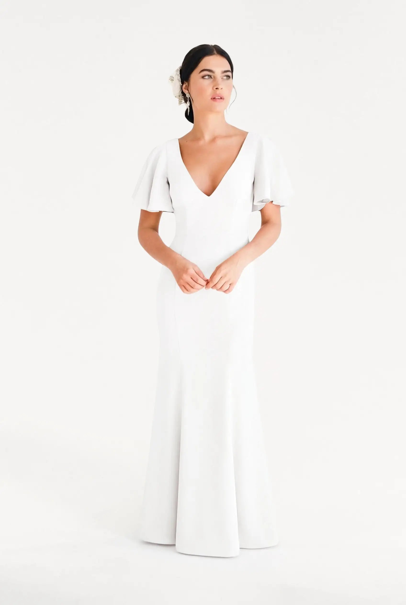 A woman with dark hair styled back and a white flower accessory wears the TH&TH Celeste Moss Crepe Dress in Ivory, a floor-length bridesmaid gown with short flutter sleeves and a deep V-neckline, standing against a plain white background.
