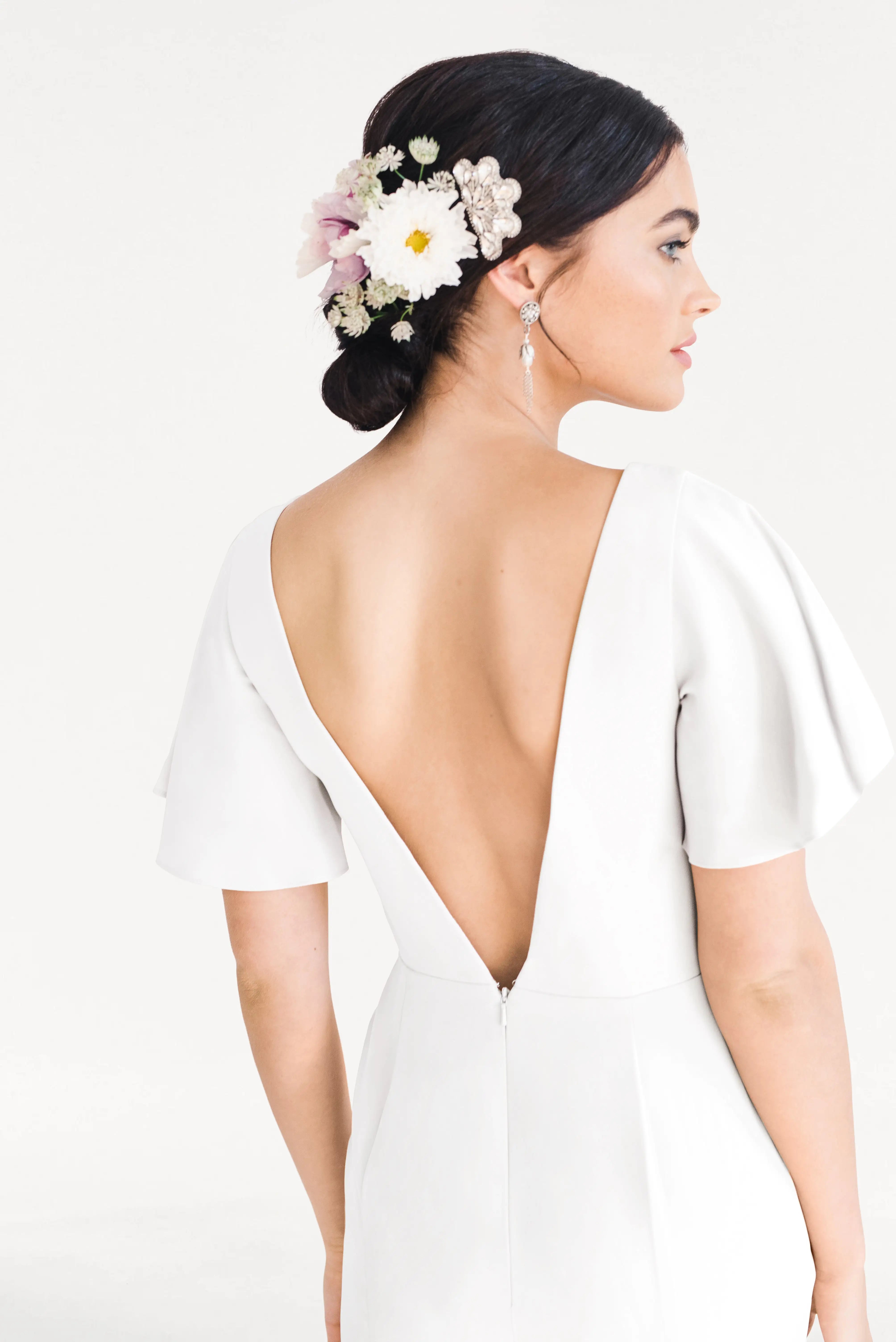 A woman with dark hair in a low bun adorned with white and pink flowers wears the TH&TH Celeste Moss Crepe Dress in Ivory, a modern, size-inclusive bridesmaid dress with short sleeves and a deep V-back, posing sideways against a plain light background.