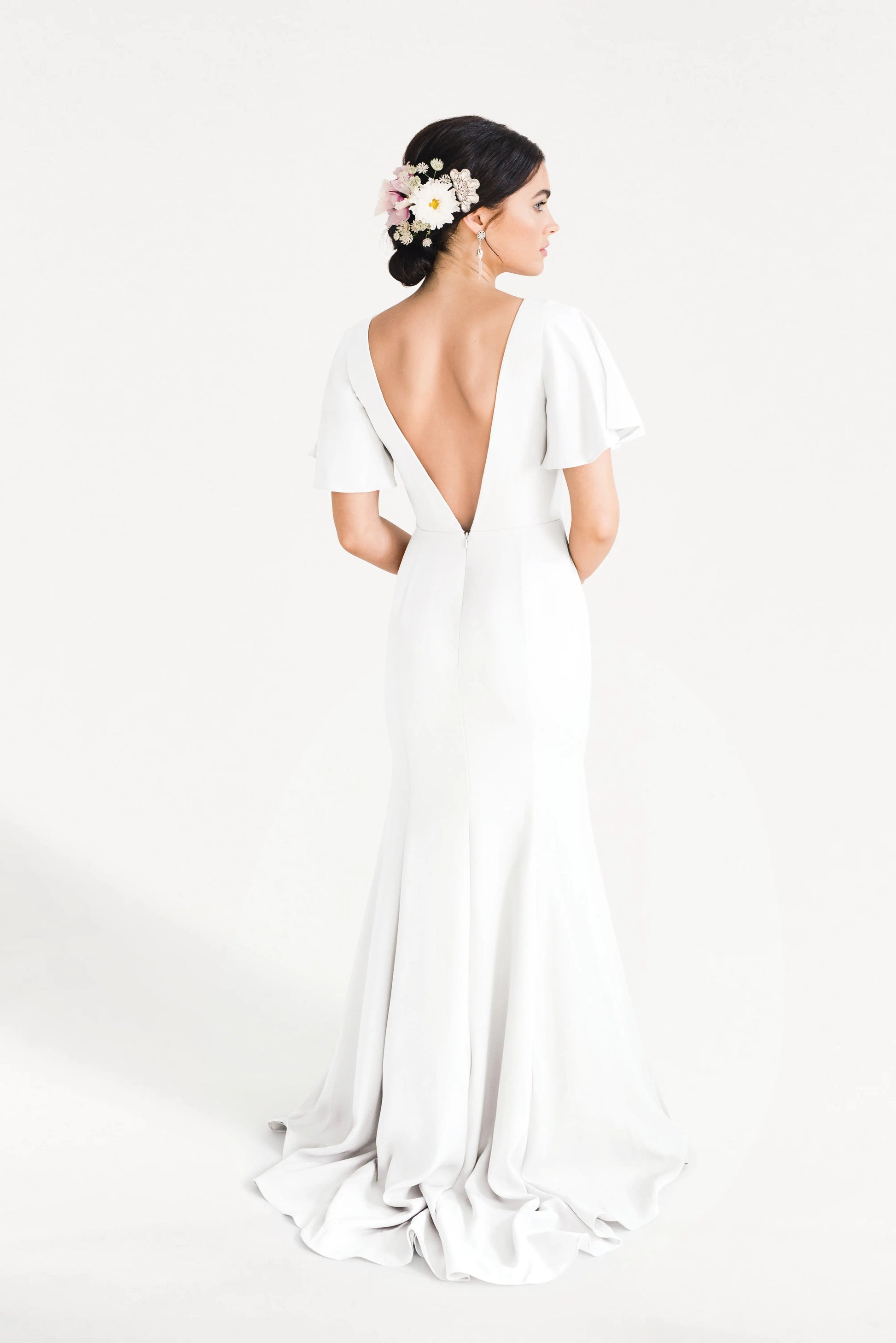 A woman stands facing away in the TH&TH Celeste Moss Crepe Dress in Ivory, a modern floor-length bridesmaid gown with a deep V-back and flutter sleeves. Her dark hair is up, adorned with pink and white flowers, against a plain white background.