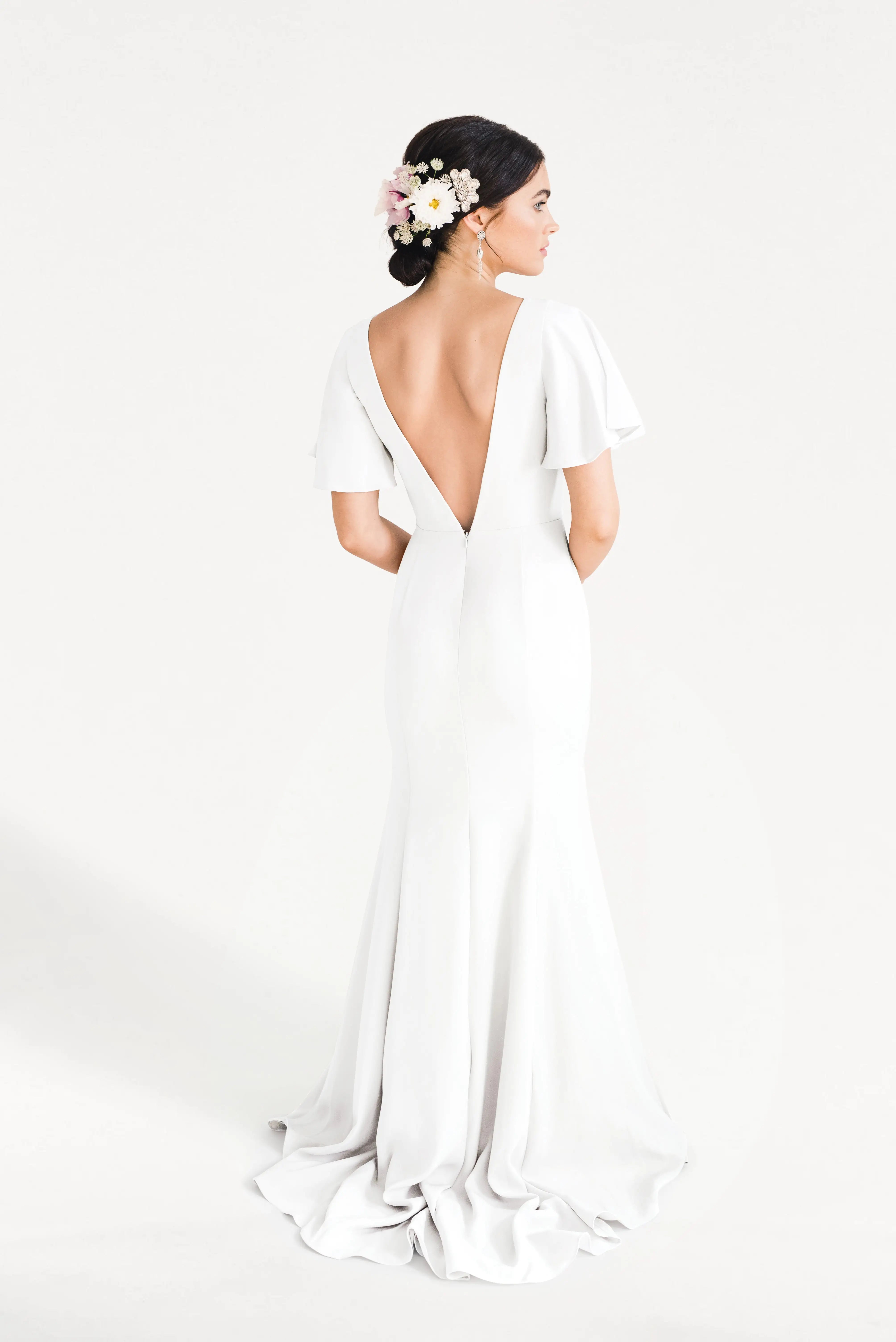A woman stands facing away in the TH&TH Celeste Moss Crepe Dress in Ivory, a modern floor-length bridesmaid gown with a deep V-back and flutter sleeves. Her dark hair is up, adorned with pink and white flowers, against a plain white background.