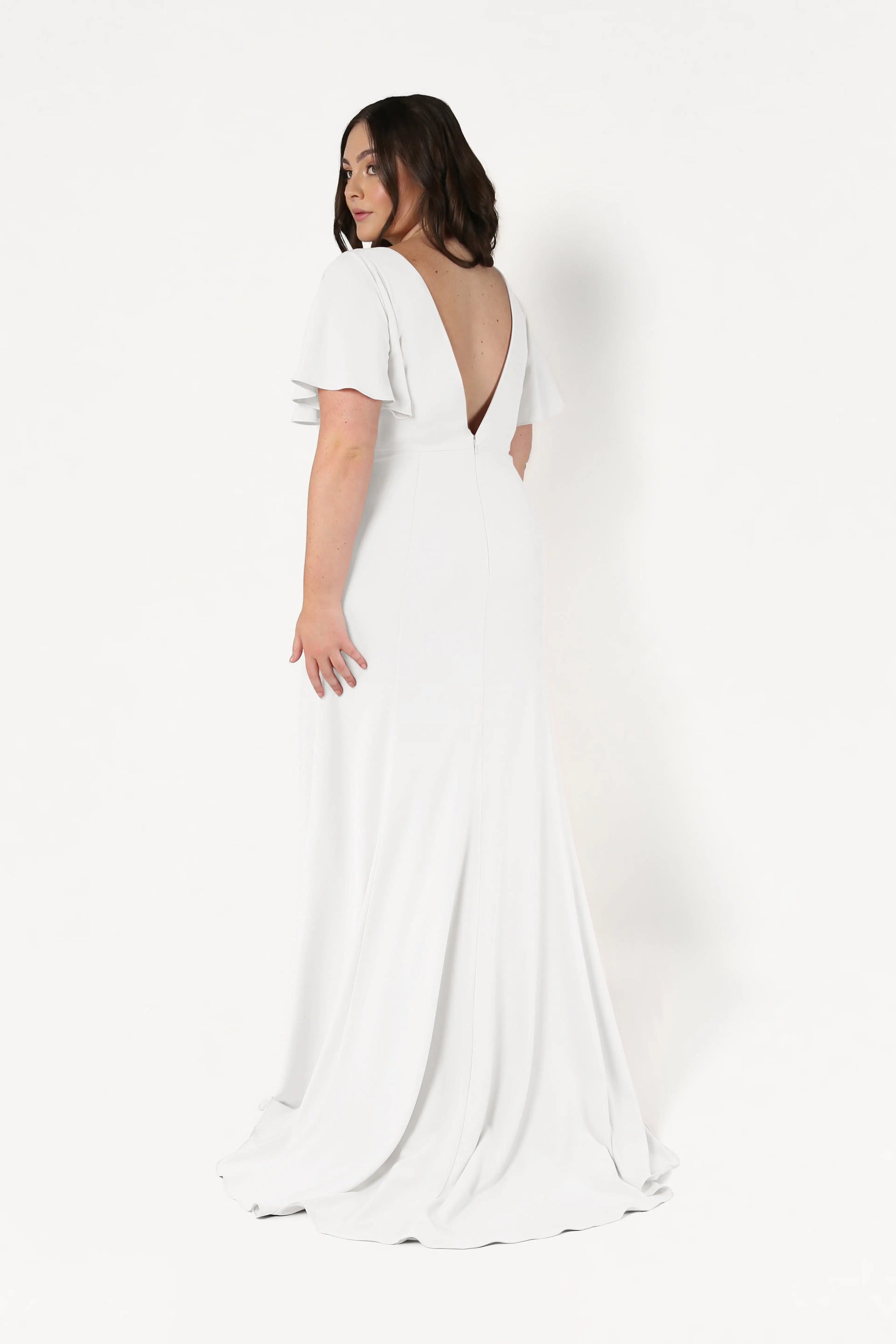 A woman stands against a plain white background, looking over her shoulder in the Celeste Moss Crepe Dress in Ivory by TH&TH—a floor-length, size-inclusive style featuring flutter sleeves and a deep V-back.