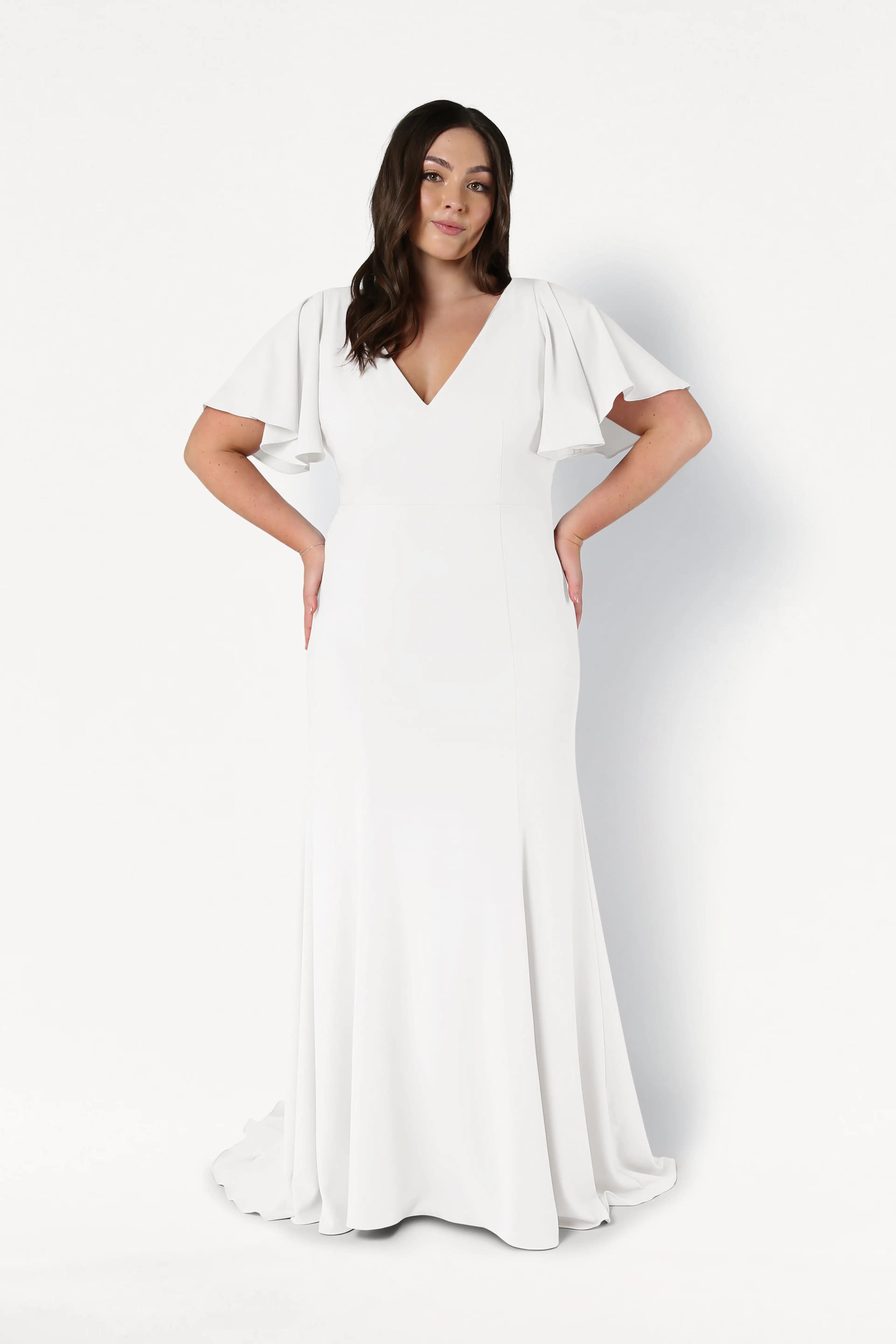A woman poses against a white background wearing the TH&TH Celeste Moss Crepe Dress in Ivory, featuring flutter sleeves and a V-neckline. Her hands are on her hips, and her hair is styled in loose waves.