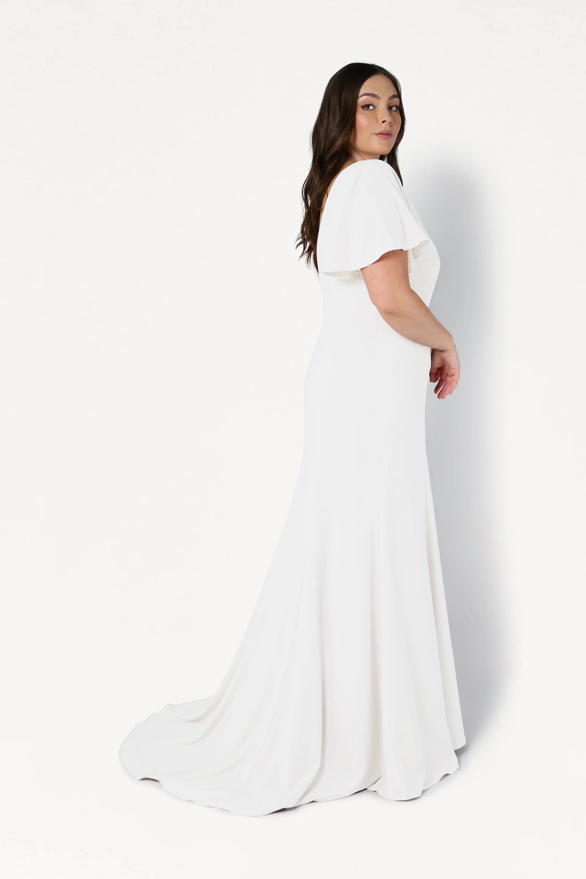 A woman models the Celeste Moss Crepe Dress in Ivory by TH&TH, featuring flutter sleeves and a flowing train, shown from a back three-quarter angle against a white background for elegant bridal style.