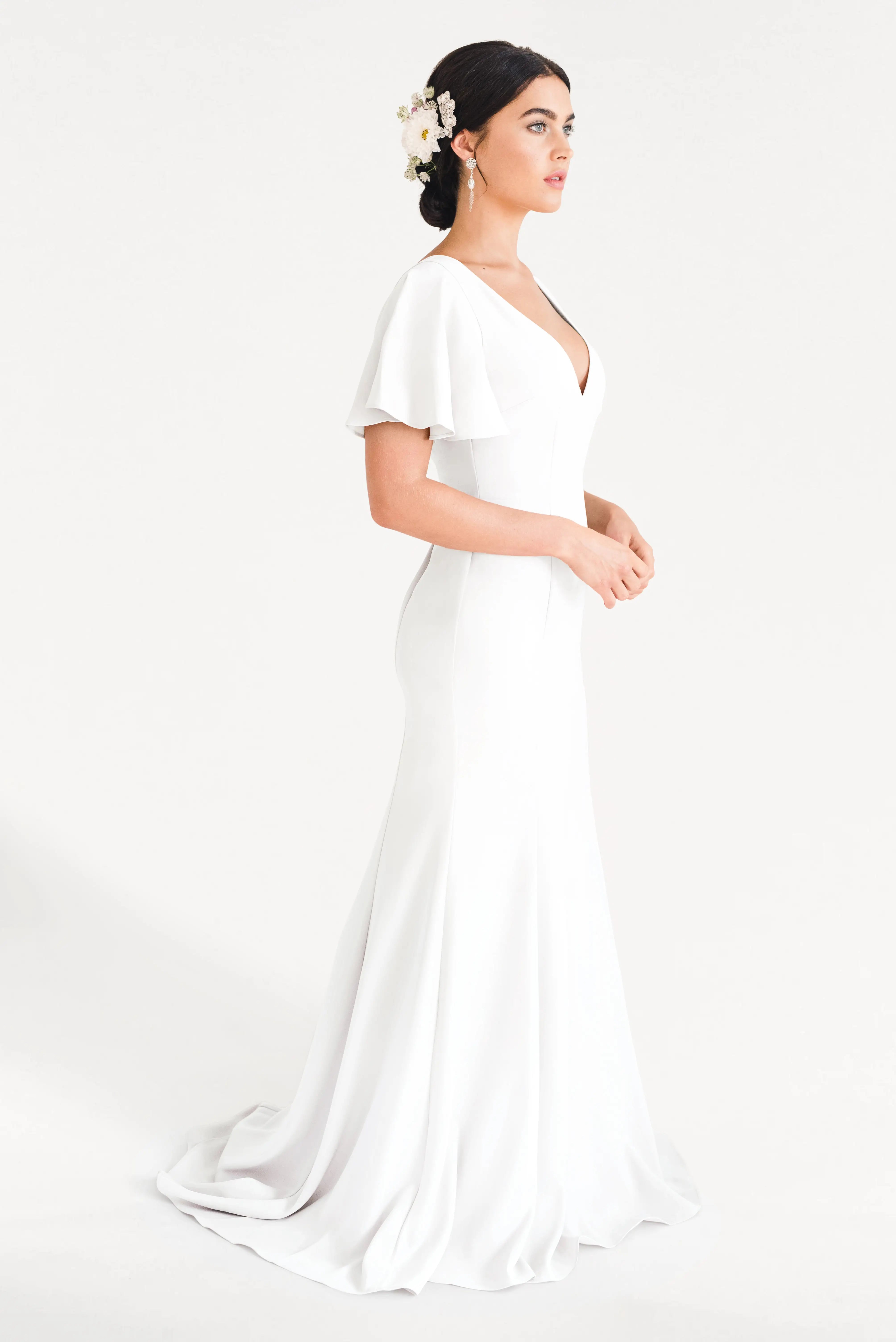 A woman wearing the TH&TH Celeste Moss Crepe Dress in Ivory, featuring flutter sleeves and a V-neckline, stands in profile with her dark updo adorned in white floral accessories against a plain white background.