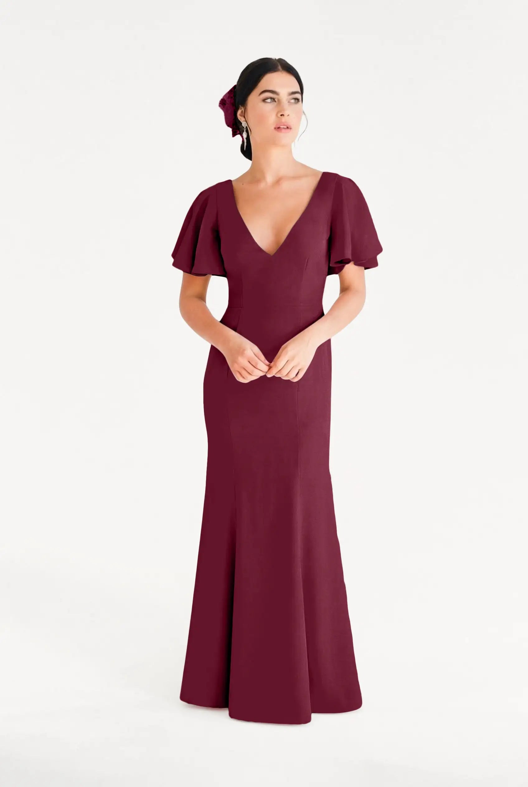A woman stands against a white background wearing the TH&TH Celeste Moss Crepe Bridesmaid Dress in Merlot, featuring short flutter sleeves, a plunging V-neckline, and a fitted, floor-length silhouette. Her dark hair is styled back with a matching accessory.