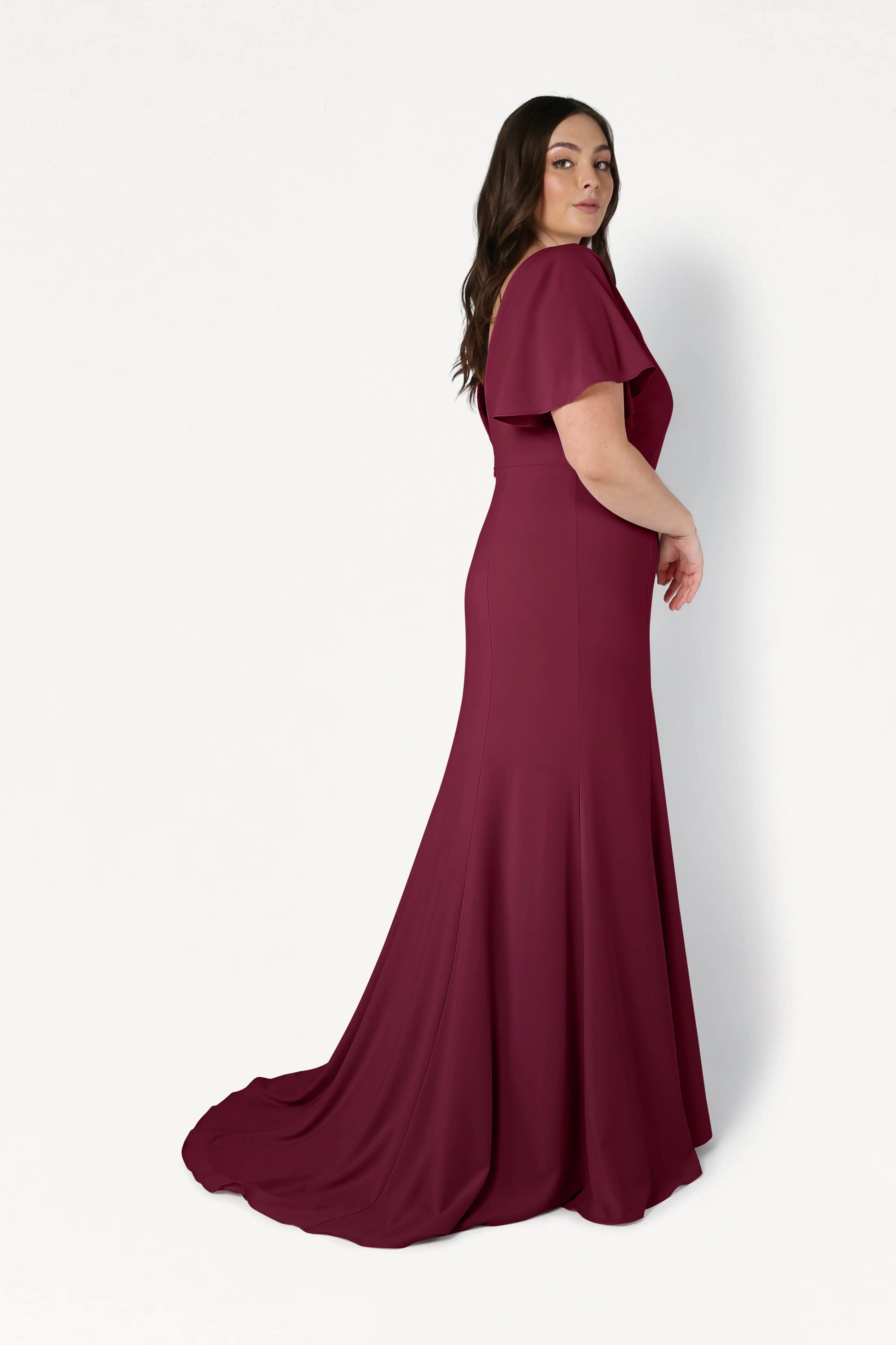A woman in the size-inclusive Celeste Moss Crepe Bridesmaid Dress in Merlot by TH&TH, featuring short flutter sleeves, a fitted bodice, and flowing train, stands in profile against a plain white background.