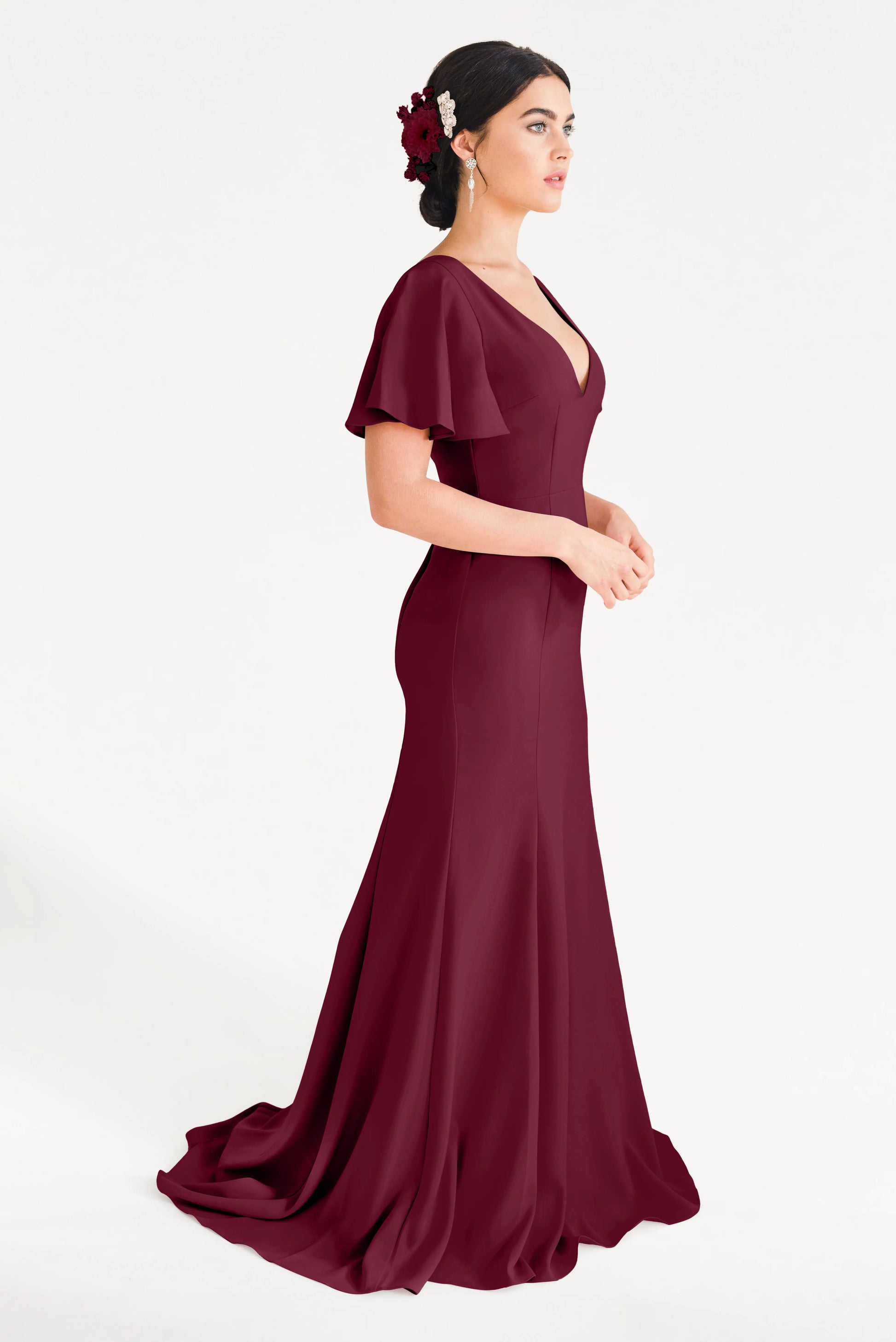 A woman in the TH&TH Celeste Moss Crepe Bridesmaid Dress in Merlot, featuring flutter sleeves and a V-neckline, stands against a white background wearing a floral hair accessory and looking to the side.