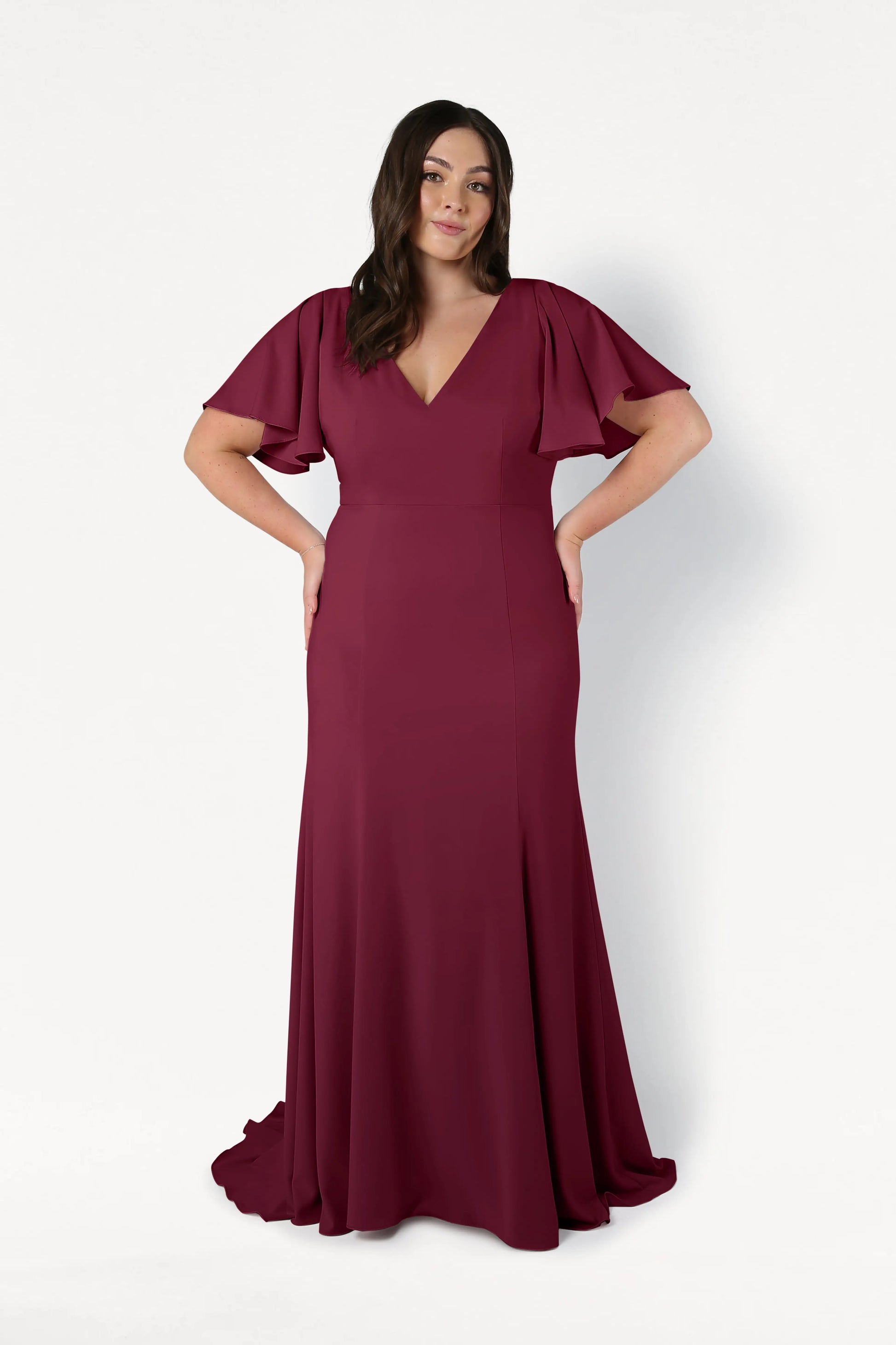 A woman stands against a white background wearing the TH&TH Celeste Moss Crepe Bridesmaid Dress in Merlot. The floor-length, size-inclusive gown features flutter sleeves, a V-neckline, and her long dark hair is styled in loose waves.