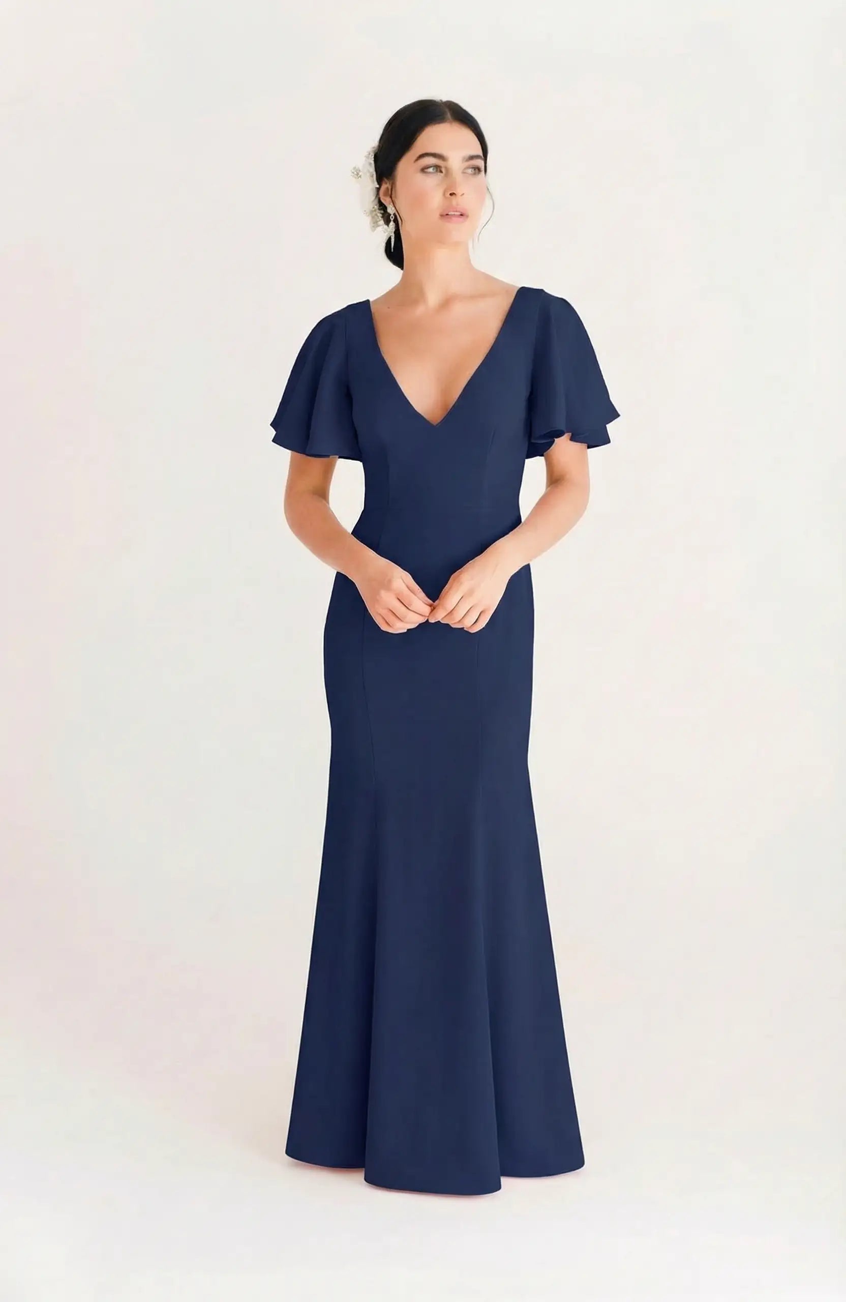A woman stands against a plain light background wearing the TH&TH Celeste Moss Crepe Bridesmaid Dress in navy, featuring short flutter sleeves, a deep V-neckline, and a floor-length silhouette.