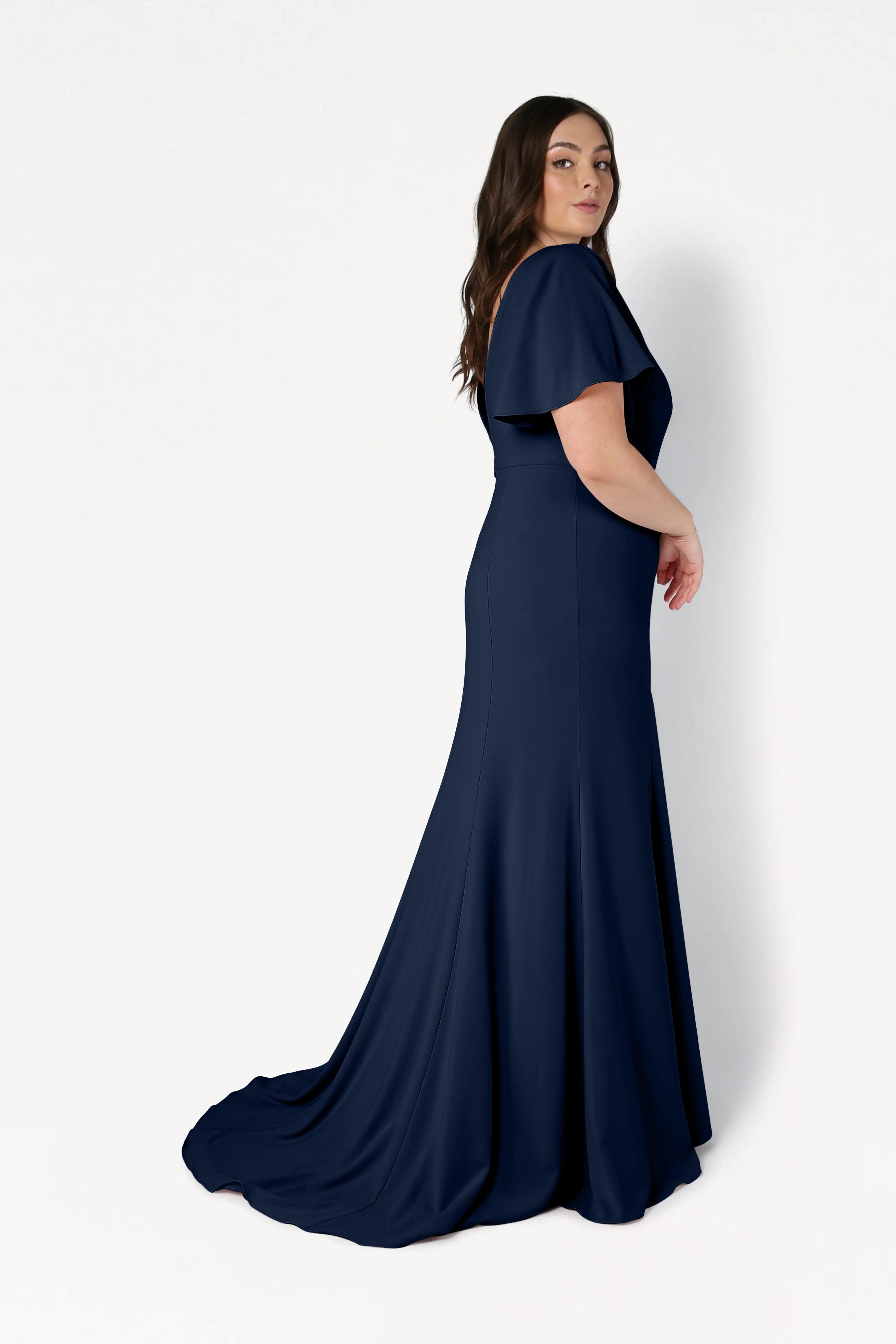 A woman with long brown hair models the floor-length Celeste Moss Crepe Bridesmaid Dress in Navy by TH&TH, featuring short sleeves and a fitted bodice, standing sideways against a plain white background.