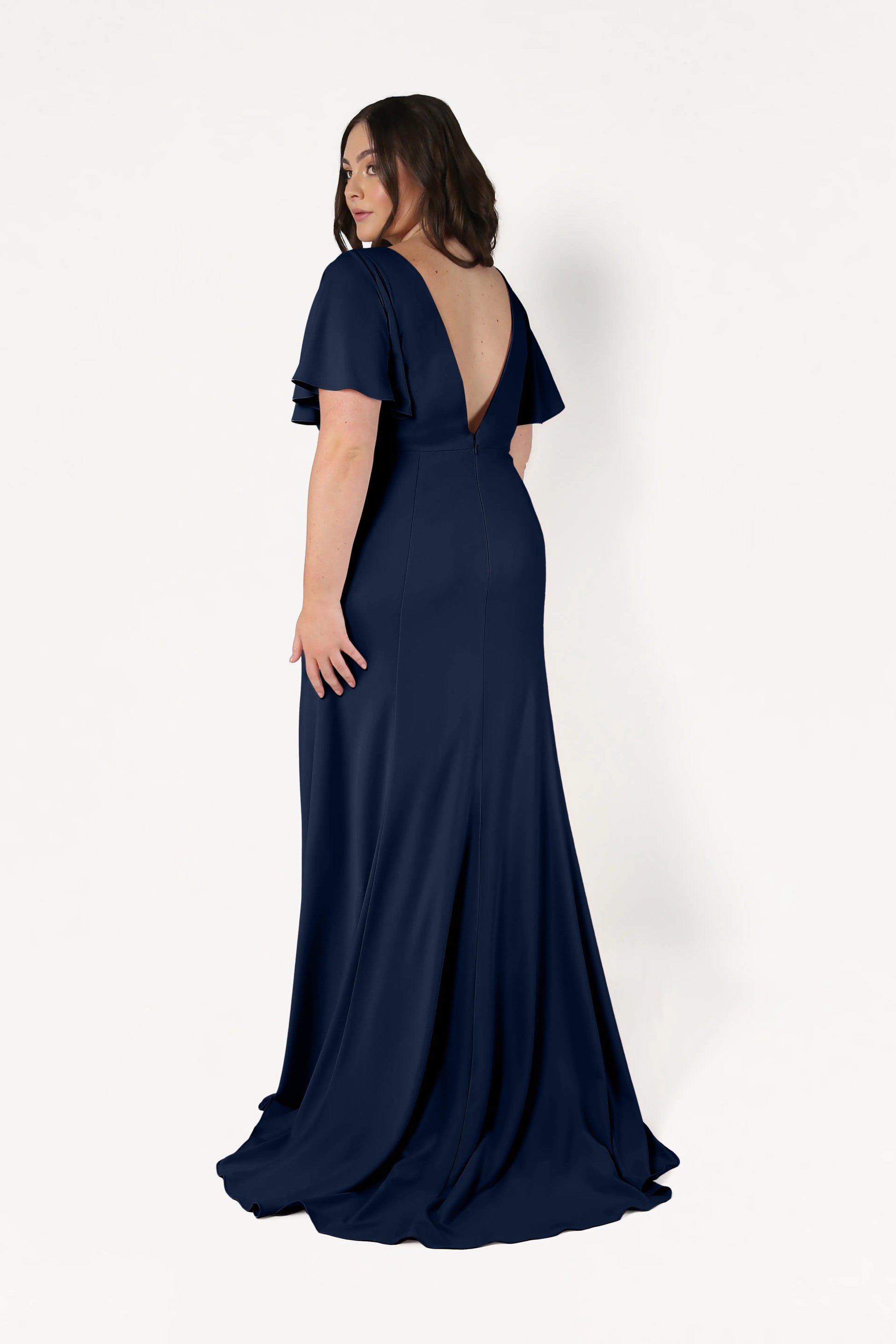 A woman in the Celeste Moss Crepe Bridesmaid Dress in Navy by TH&TH, featuring a floor-length silhouette, short flutter sleeves, and a deep V-back, stands against a white background and looks over her shoulder.