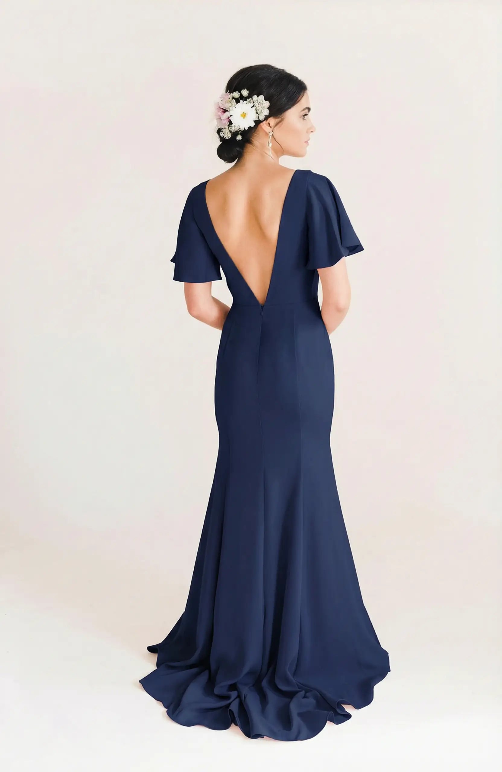 A woman with dark hair in a low bun adorned with flowers wears the TH&TH Celeste Moss Crepe Bridesmaid Dress in Navy, featuring flutter sleeves and a deep V-back, standing against a plain light background.