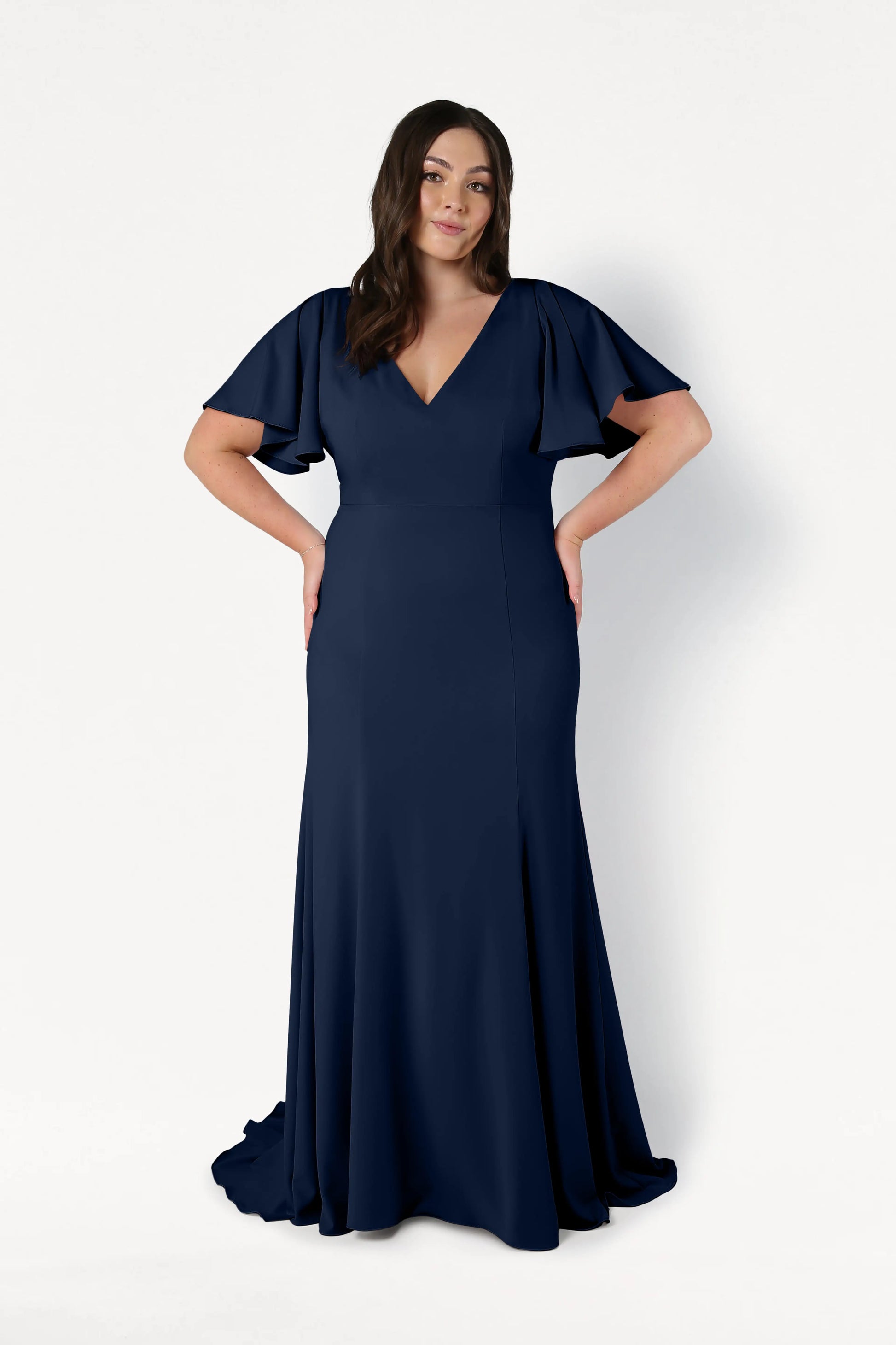 A woman models the Celeste Moss Crepe Bridesmaid Dress in Navy by TH&TH, featuring flutter sleeves and a V-neckline; she stands against a white background with her hands on her hips.