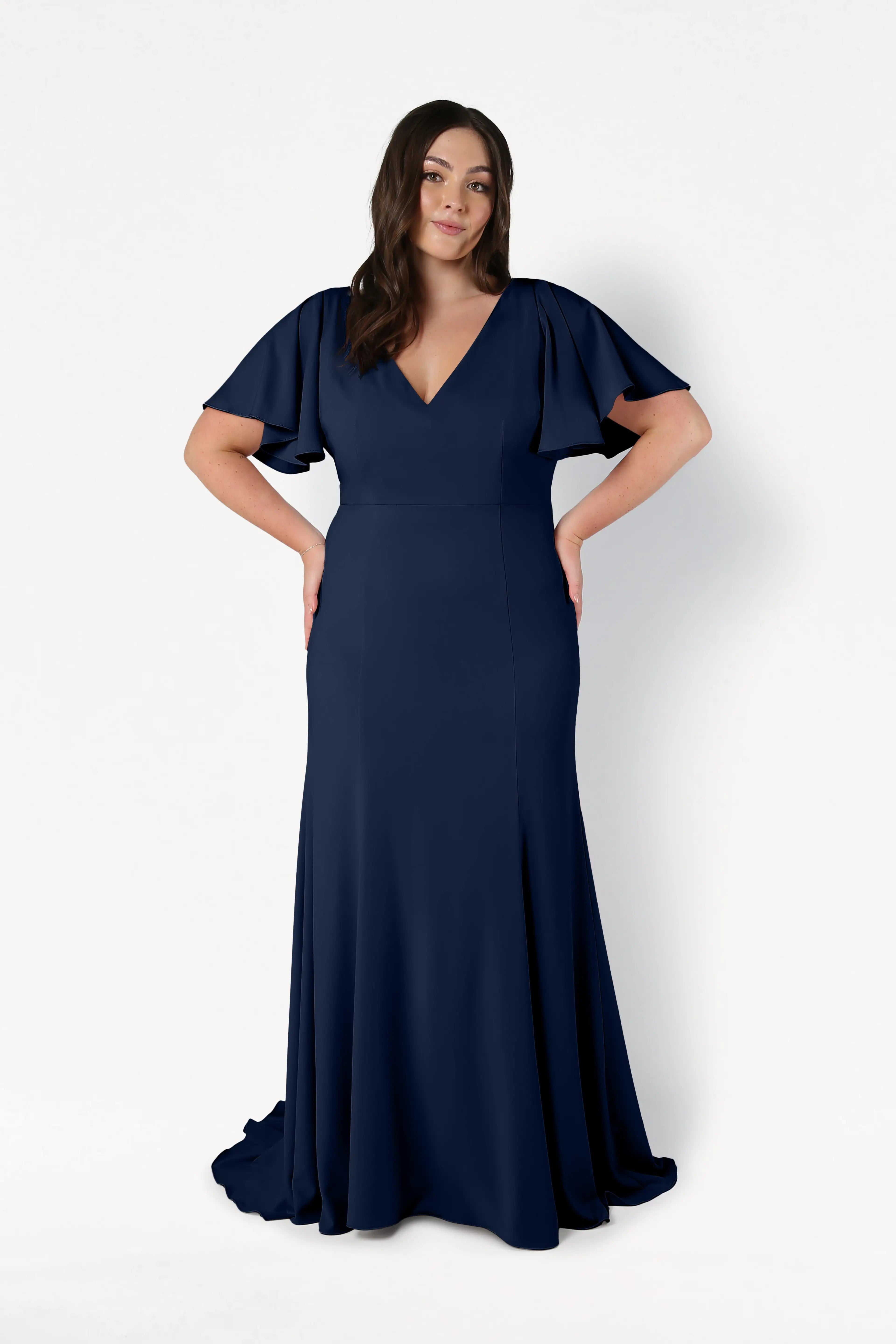 A woman models the Celeste Moss Crepe Bridesmaid Dress in Navy by TH&TH, featuring flutter sleeves and a V-neckline; she stands against a white background with her hands on her hips.