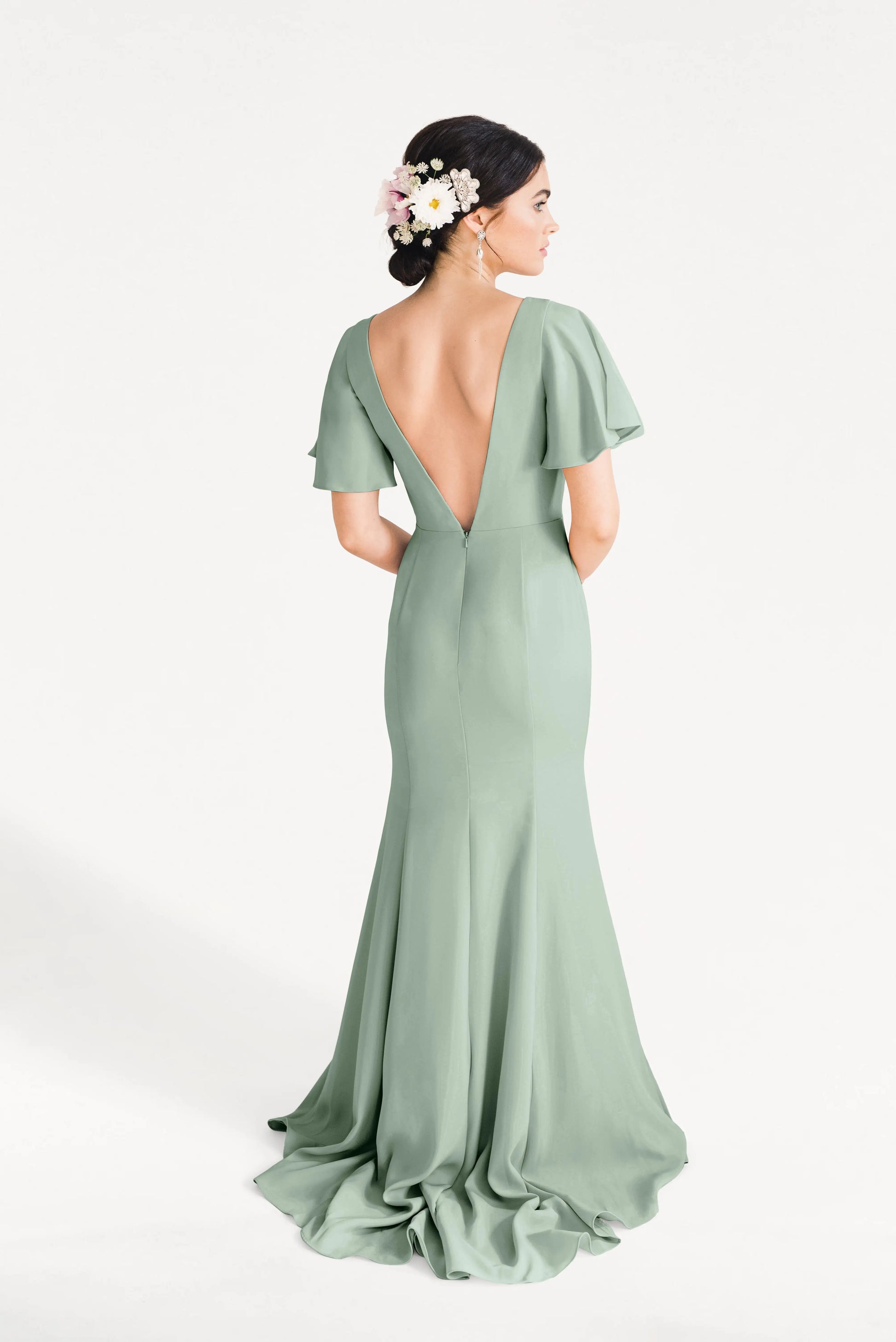 A woman with dark hair in a floral bun models the Celeste Moss Crepe Bridesmaid Dress in Sage Green by TH&TH—a size-inclusive gown featuring flutter sleeves and a deep V-back—against a white background.