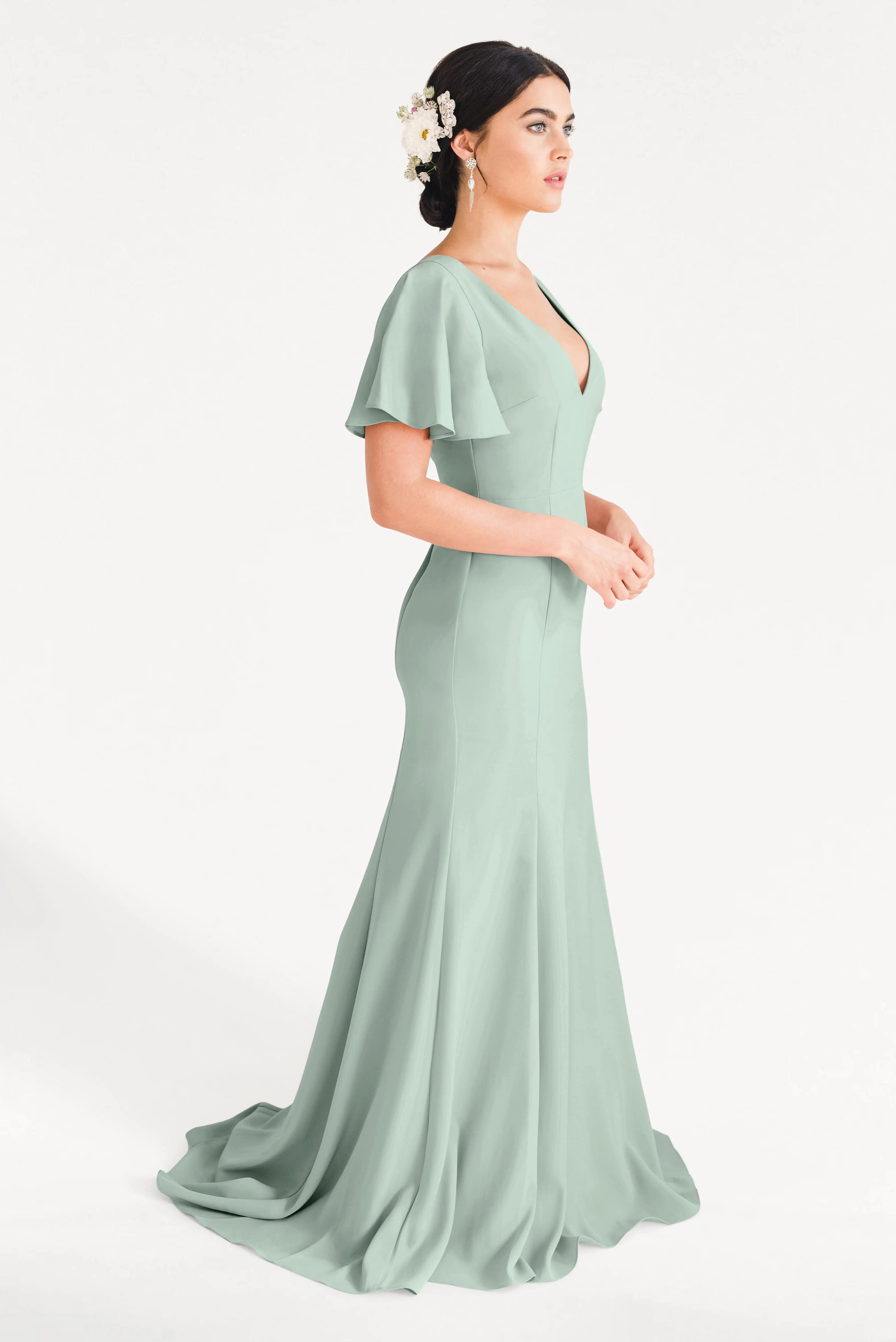A woman in the TH&TH Celeste Moss Crepe Bridesmaid Dress in Sage Green, a floor-length gown with flutter sleeves, stands in profile. Her dark hair is in an updo with white floral accents and she wears drop earrings against a plain white background.