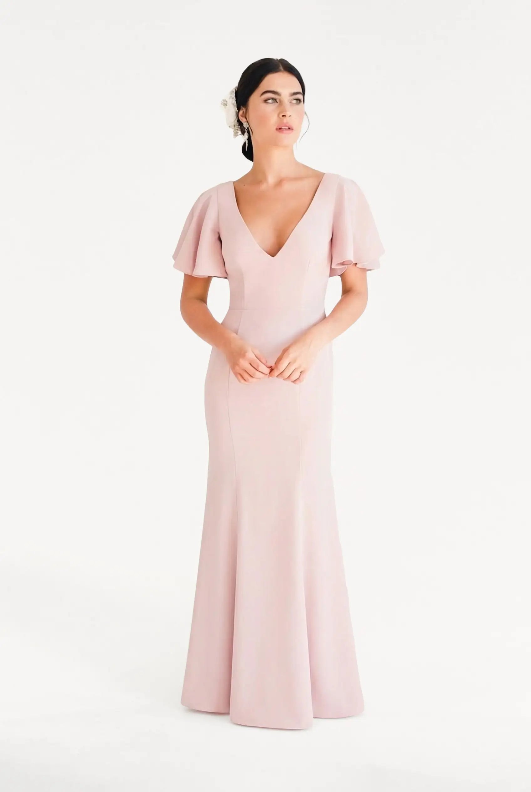 A woman models the TH&TH Celeste Moss Crepe Bridesmaid Dress in Smoked Blush, a size-inclusive, floor-length gown with short flutter sleeves and a deep V-neckline. She wears her dark hair back with a white flower accessory.